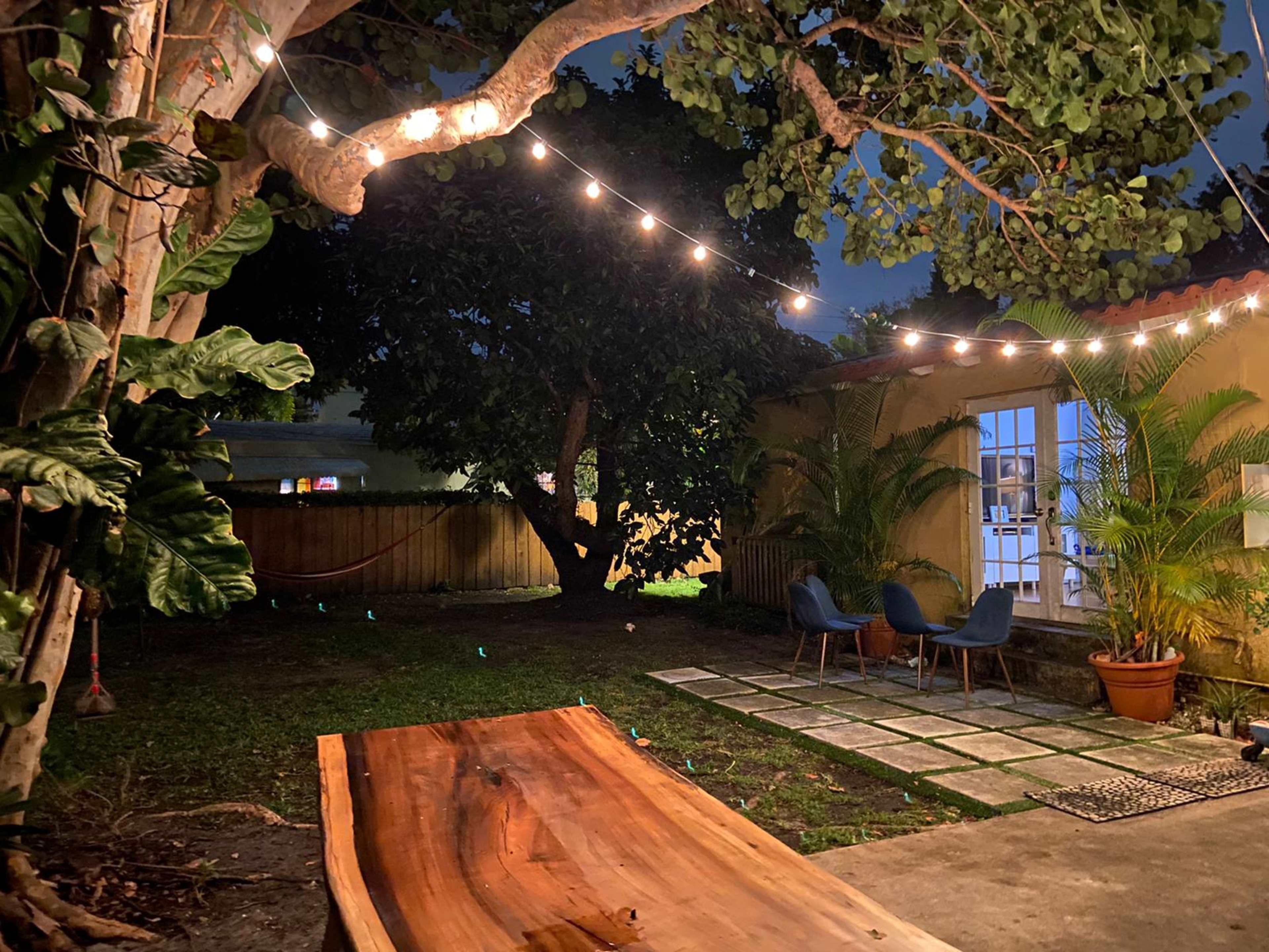 A backyard at night, featuring string lights overhead, a wooden table in the foreground, and two chairs beside a small garden area.