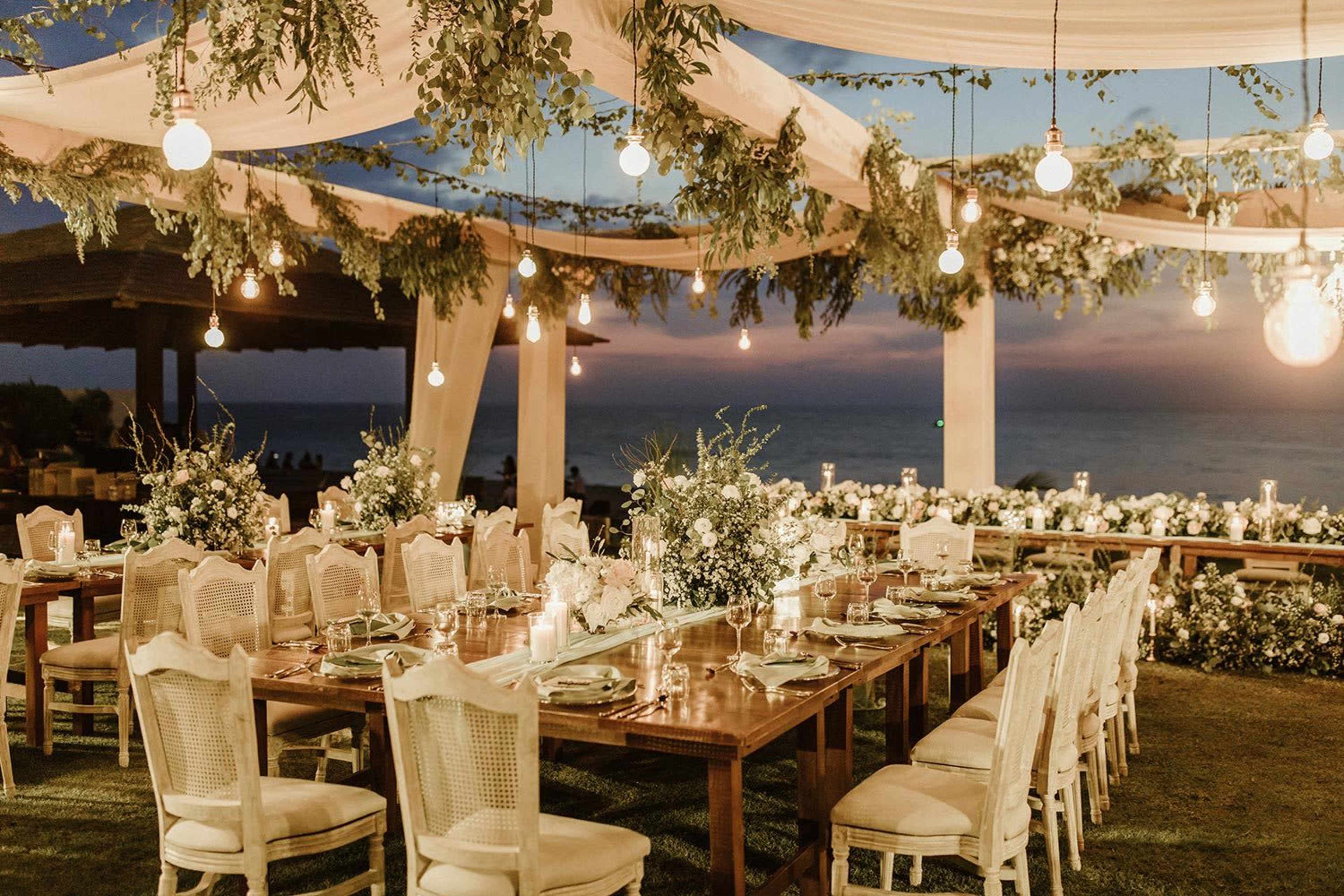 A beautifully arranged outdoor dining setup features long wooden tables adorned with floral centerpieces and suspended lights, overlooking the ocean at sunset.