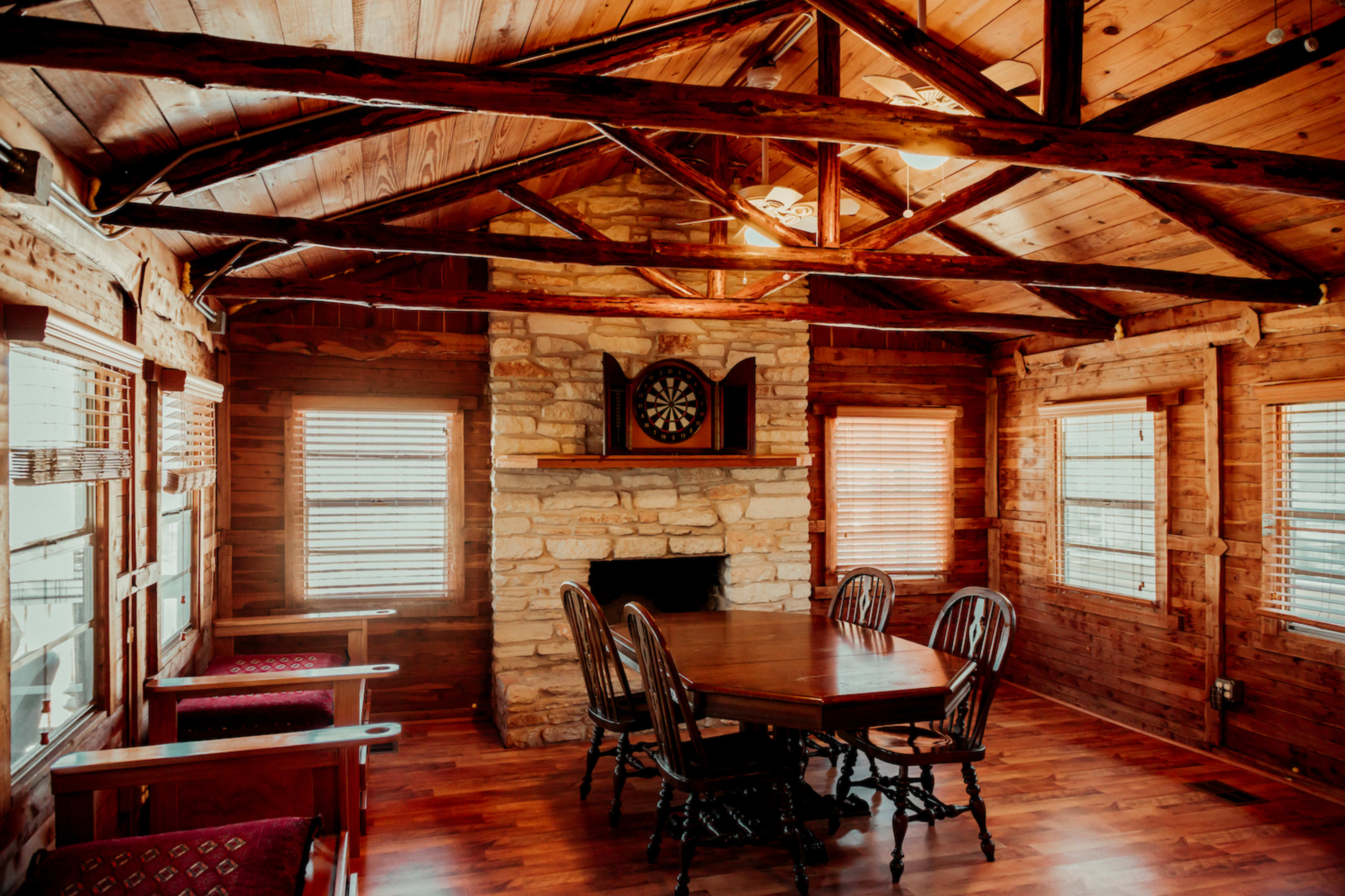 A rustic interior of a cabin with wooden walls and ceiling beams, featuring a stone fireplace, a round dining table, and several chairs.
