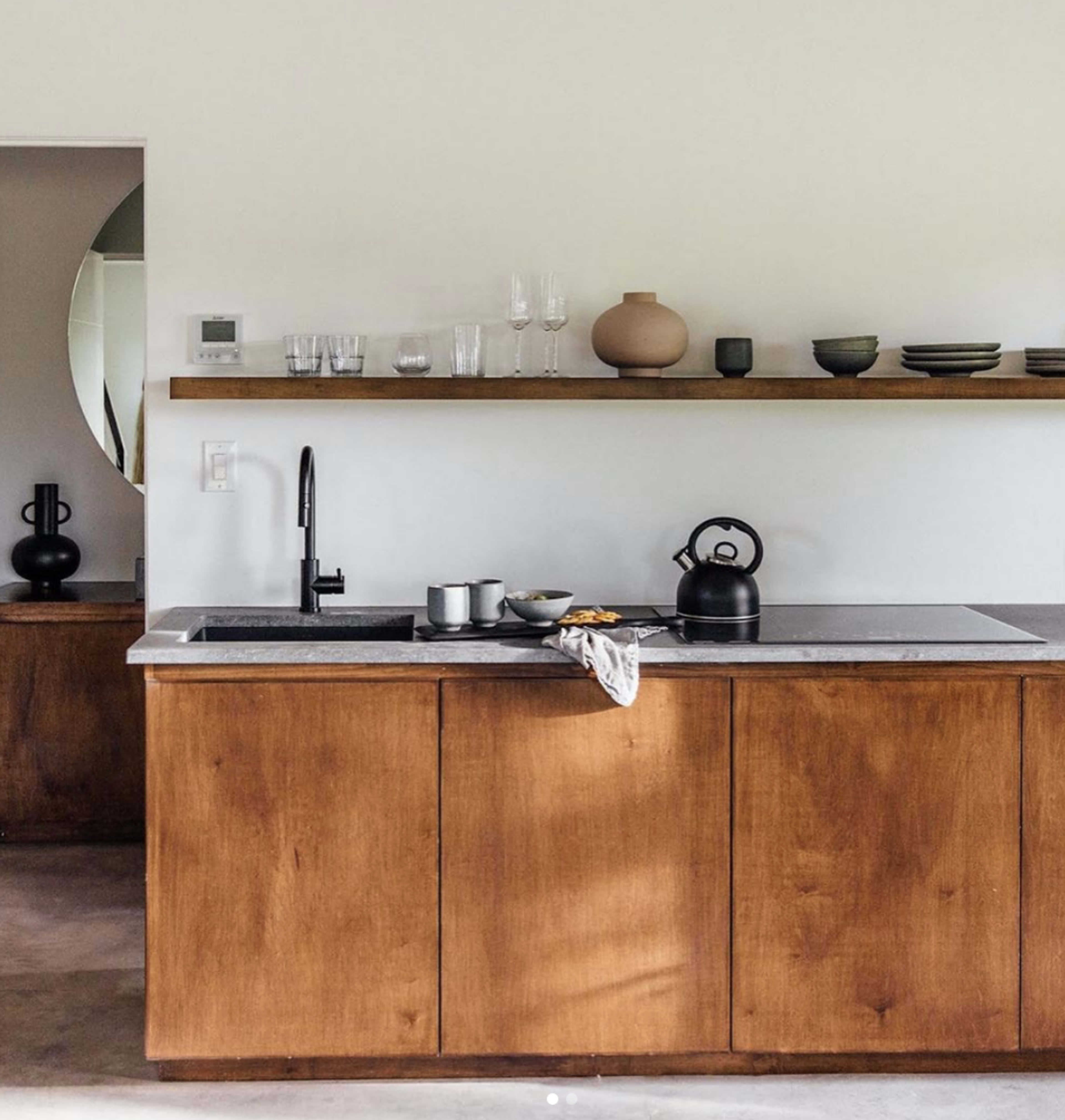 The image shows a modern kitchen with wooden cabinetry, a gray countertop, and minimalist decor on a floating shelf.