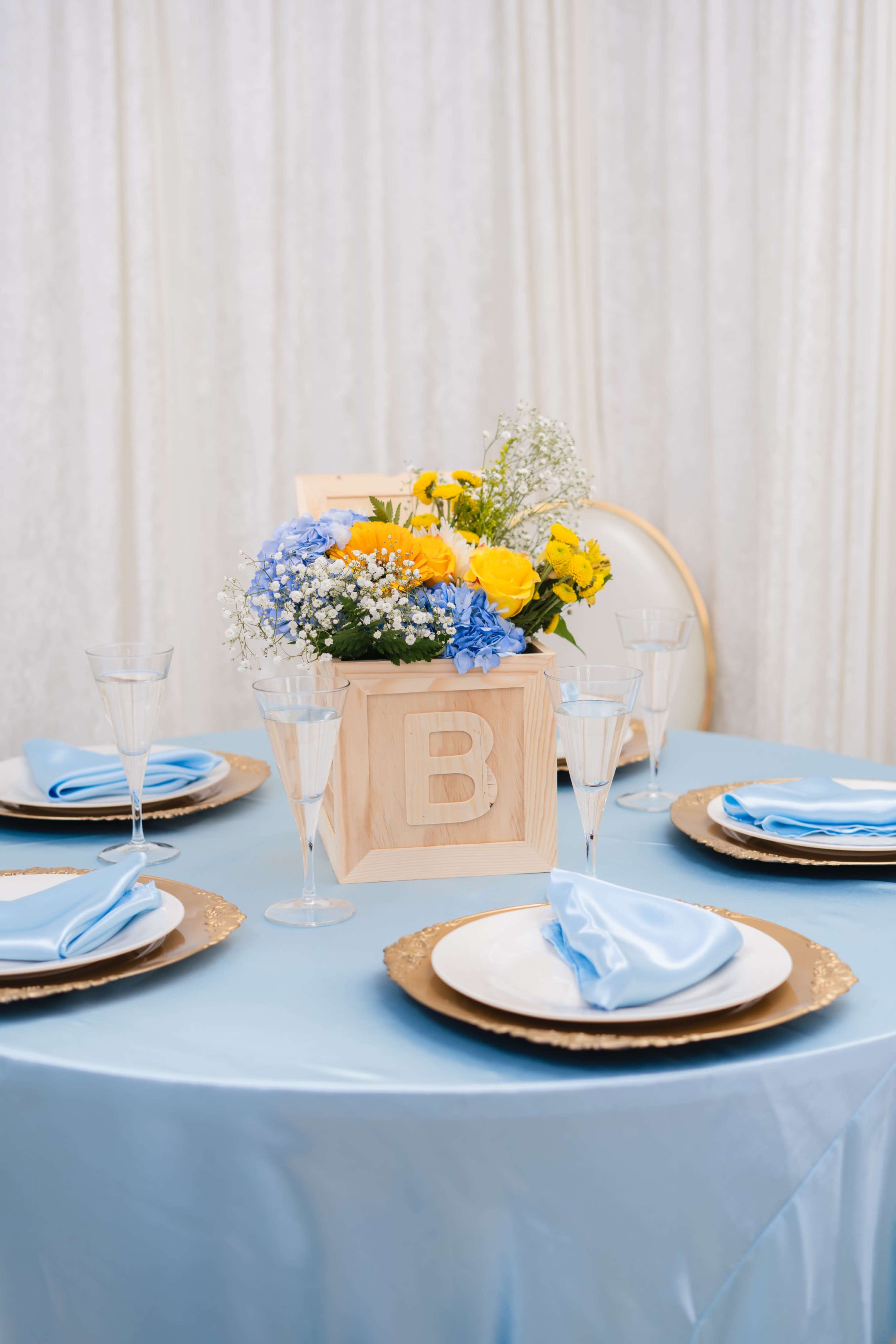 A table is set with blue tablecloth, decorative plates, and a wooden box of flowers in the center.