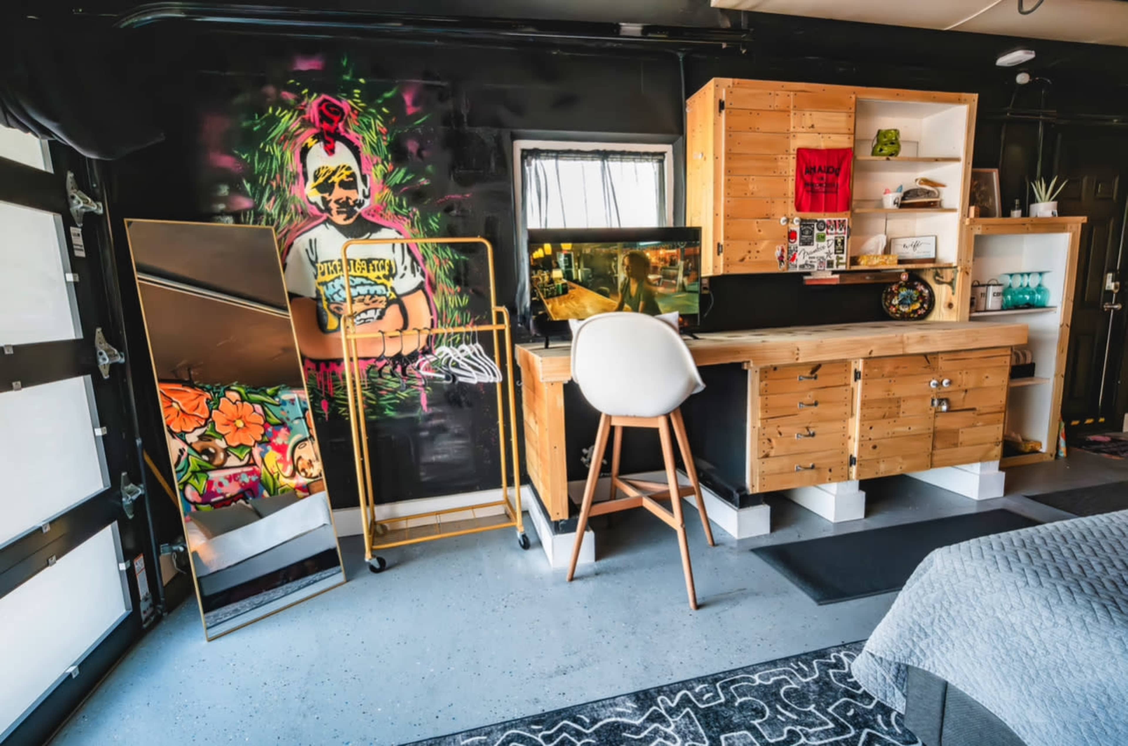 The image shows a modern, industrial-themed room featuring a wooden desk and storage, a large mirror, and a wall adorned with colorful street art.
