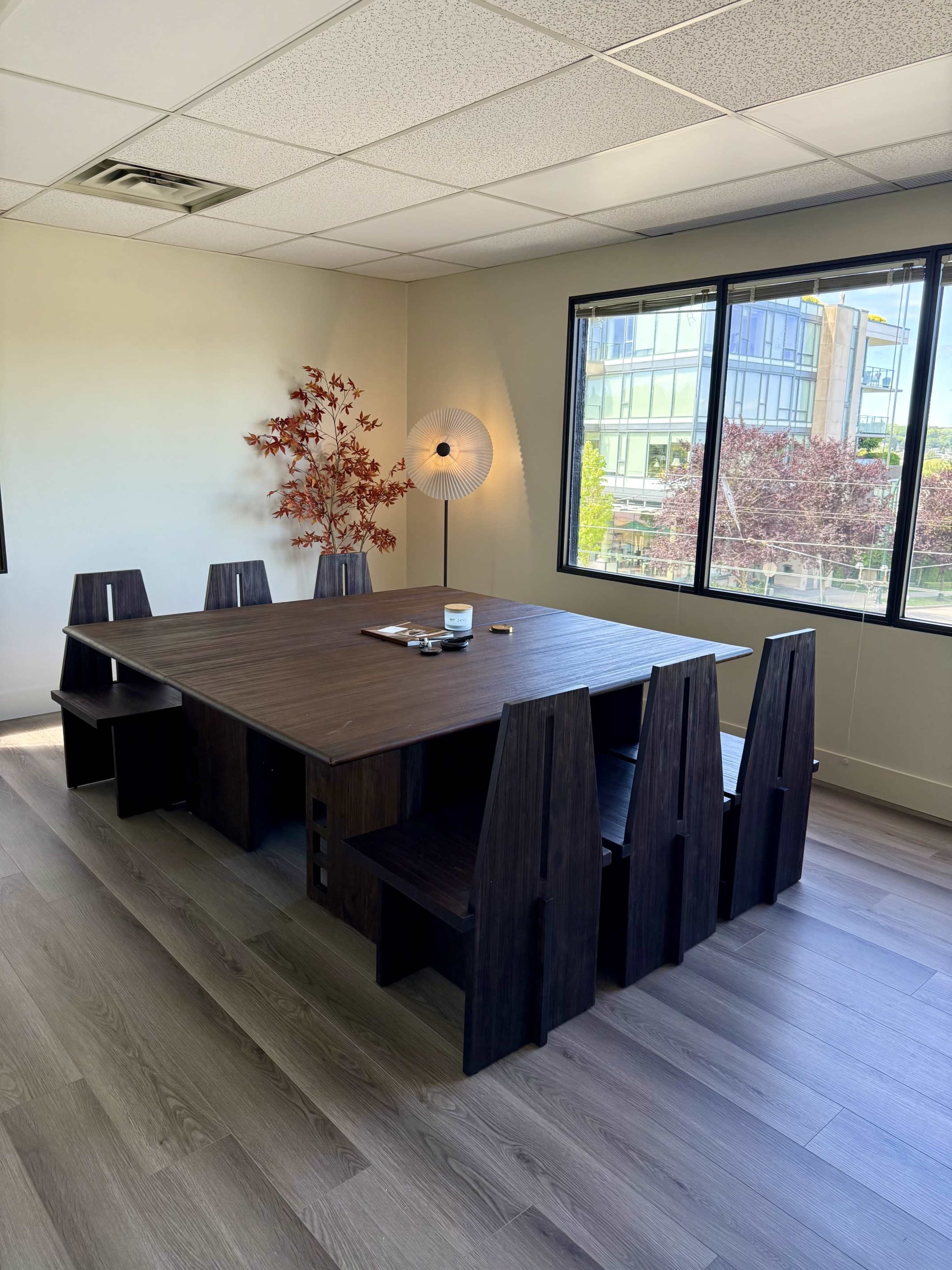 A minimalist conference room features a large wooden table surrounded by six geometric chairs, with a decorative plant and large windows offering outdoor views.