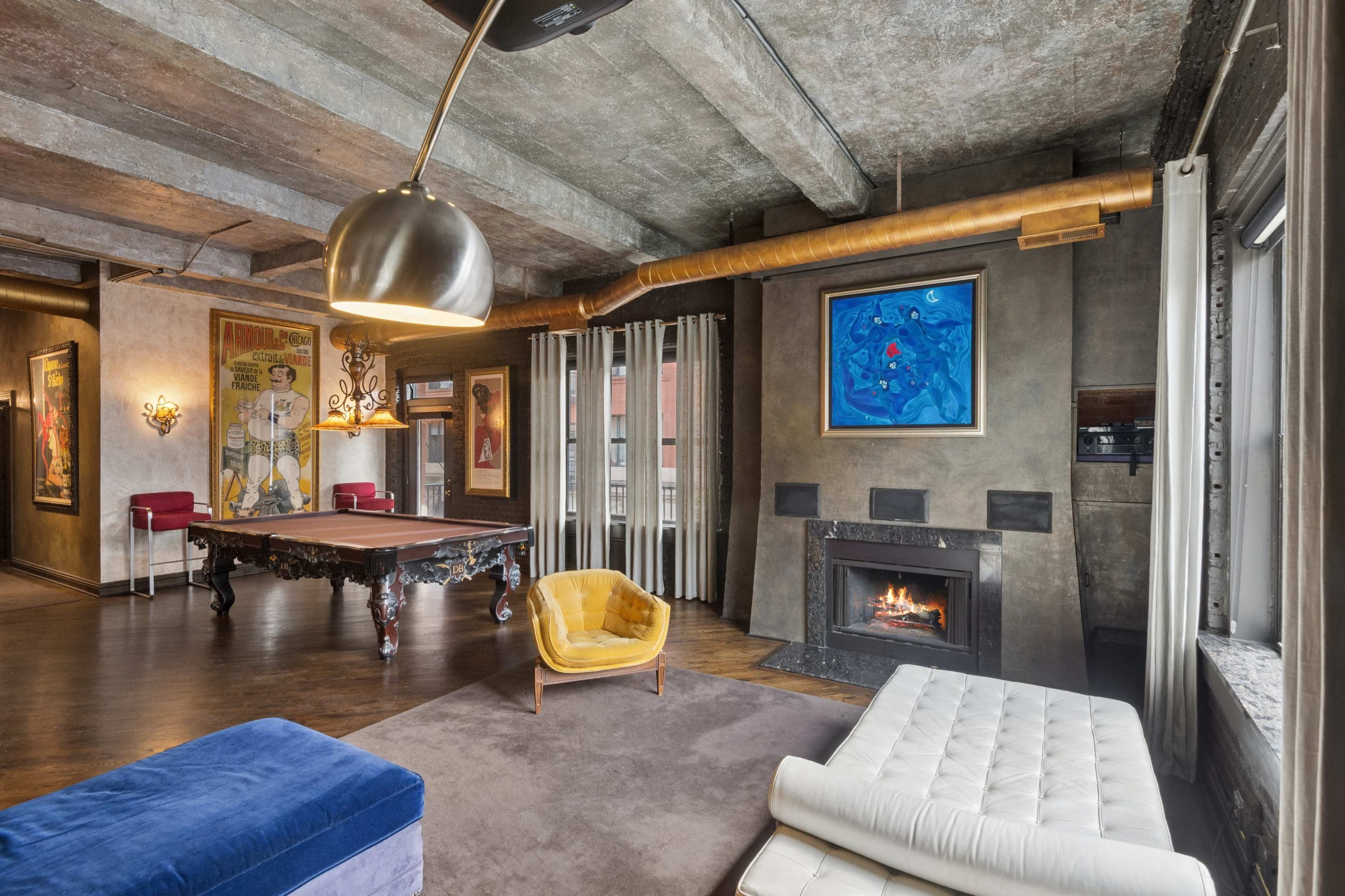 The image shows a modern living space featuring a pool table, a fireplace, and stylish furniture set against exposed concrete walls and large windows.
