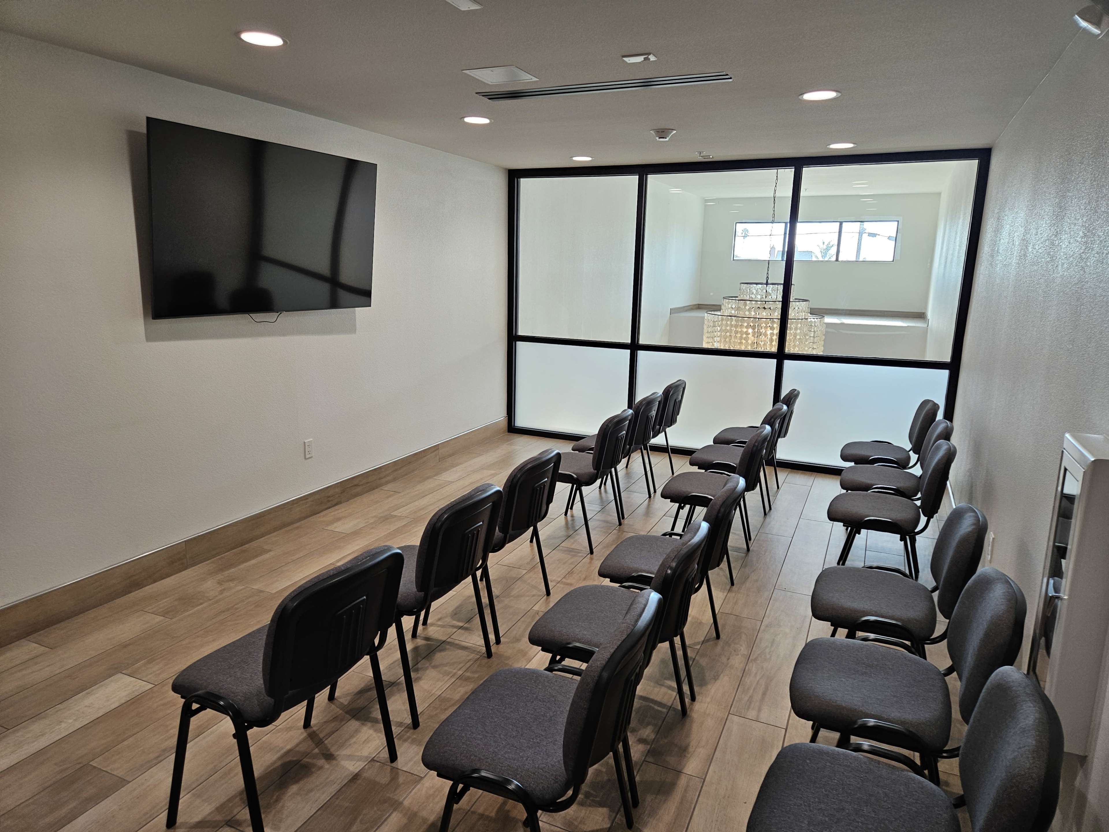 A meeting room with a television mounted on the wall, facing a row of chairs arranged for seating.
