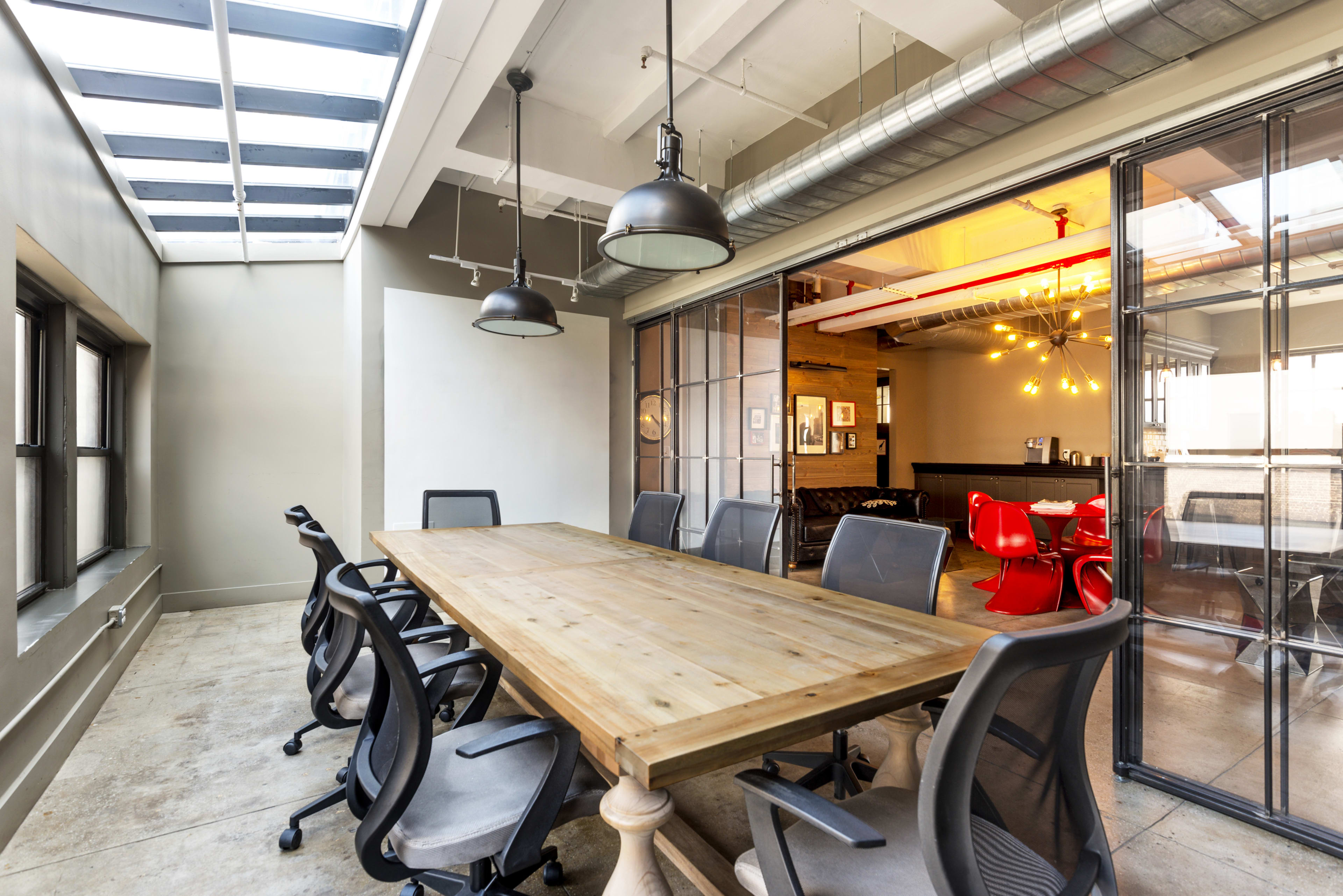 A large wooden conference table with black office chairs is placed in a well-lit meeting room featuring glass walls and pendant lights.
