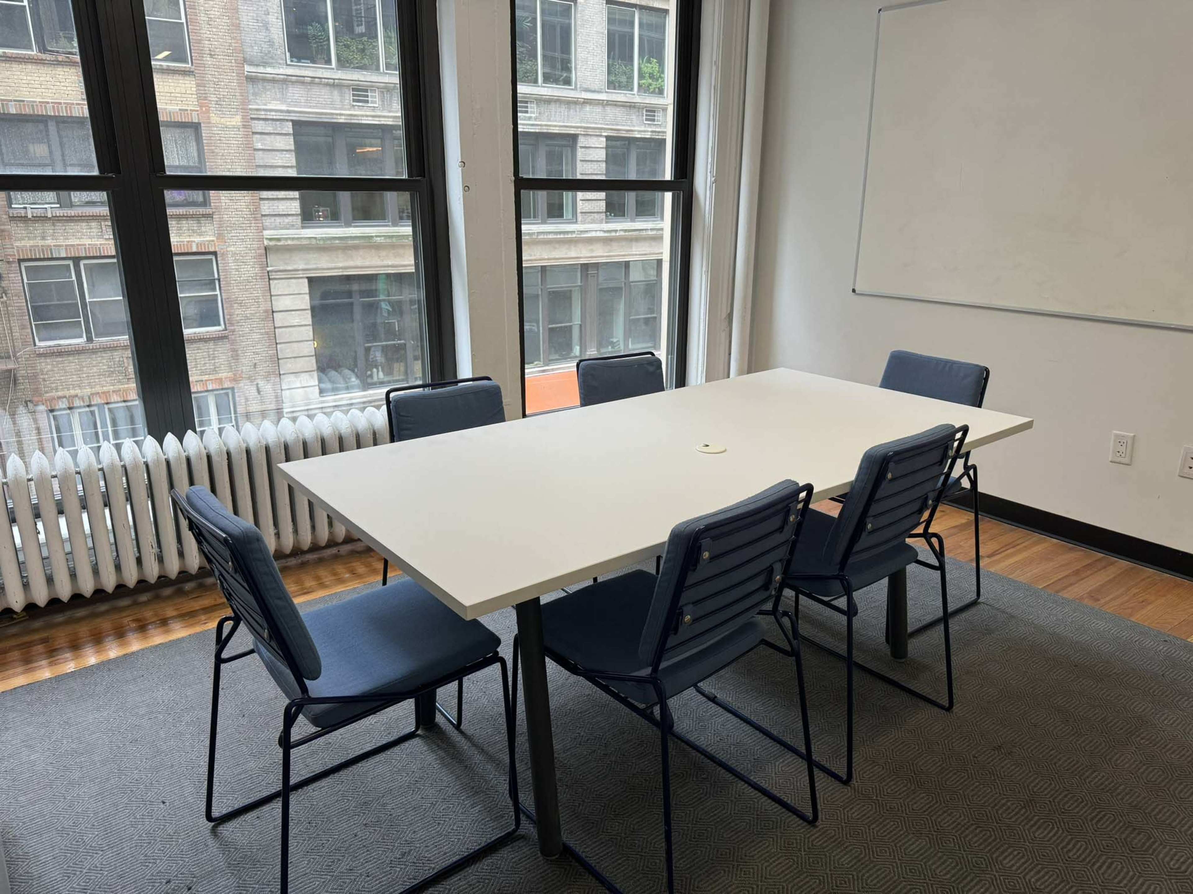 A conference room features a large white table surrounded by six chairs, with windows overlooking a city street.