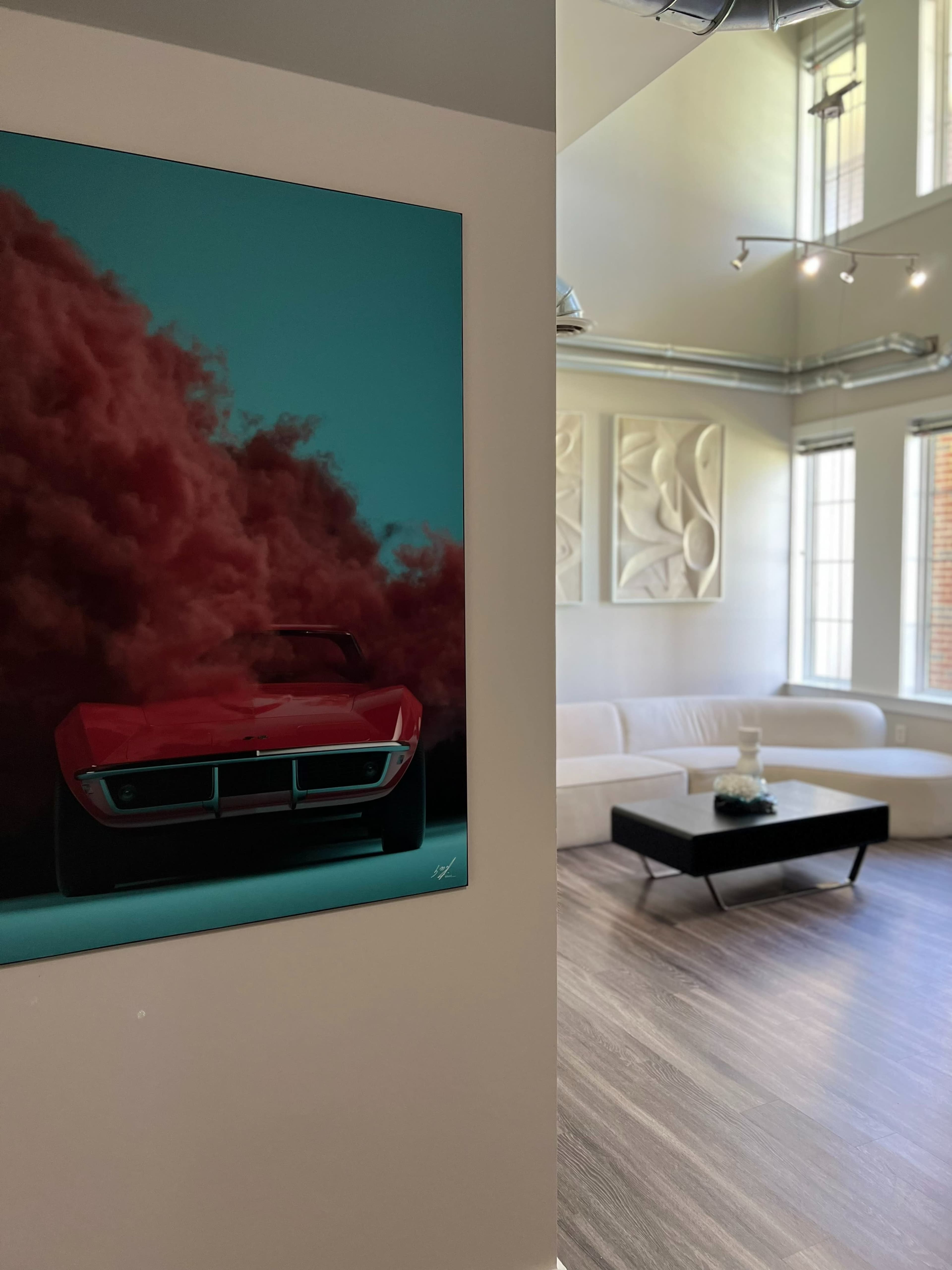 A red sports car painting hangs on a wall beside a modern living room with a white sofa and a black coffee table.
