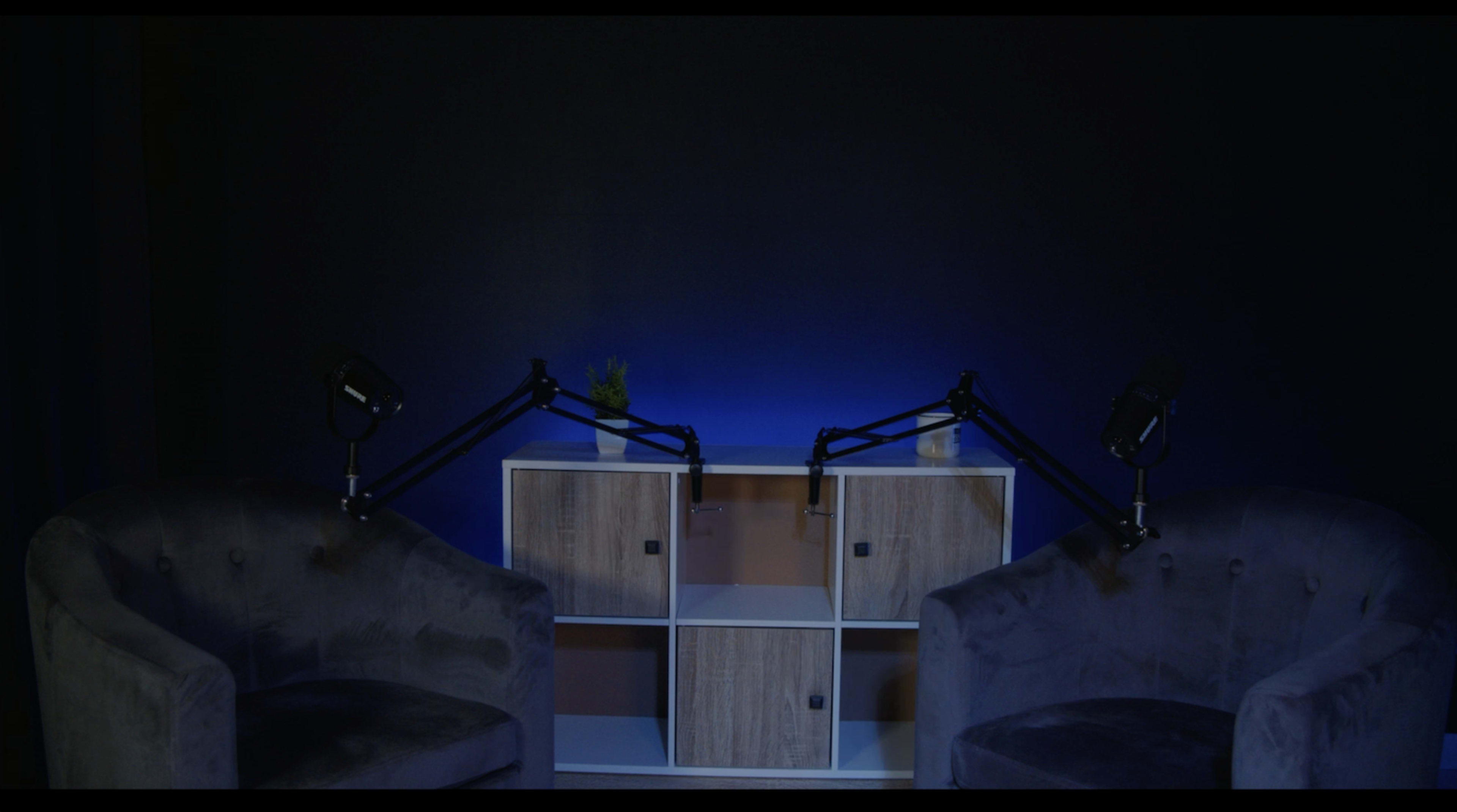Two dark, plush armchairs are positioned facing each other in front of a shelf with blue lighting.
