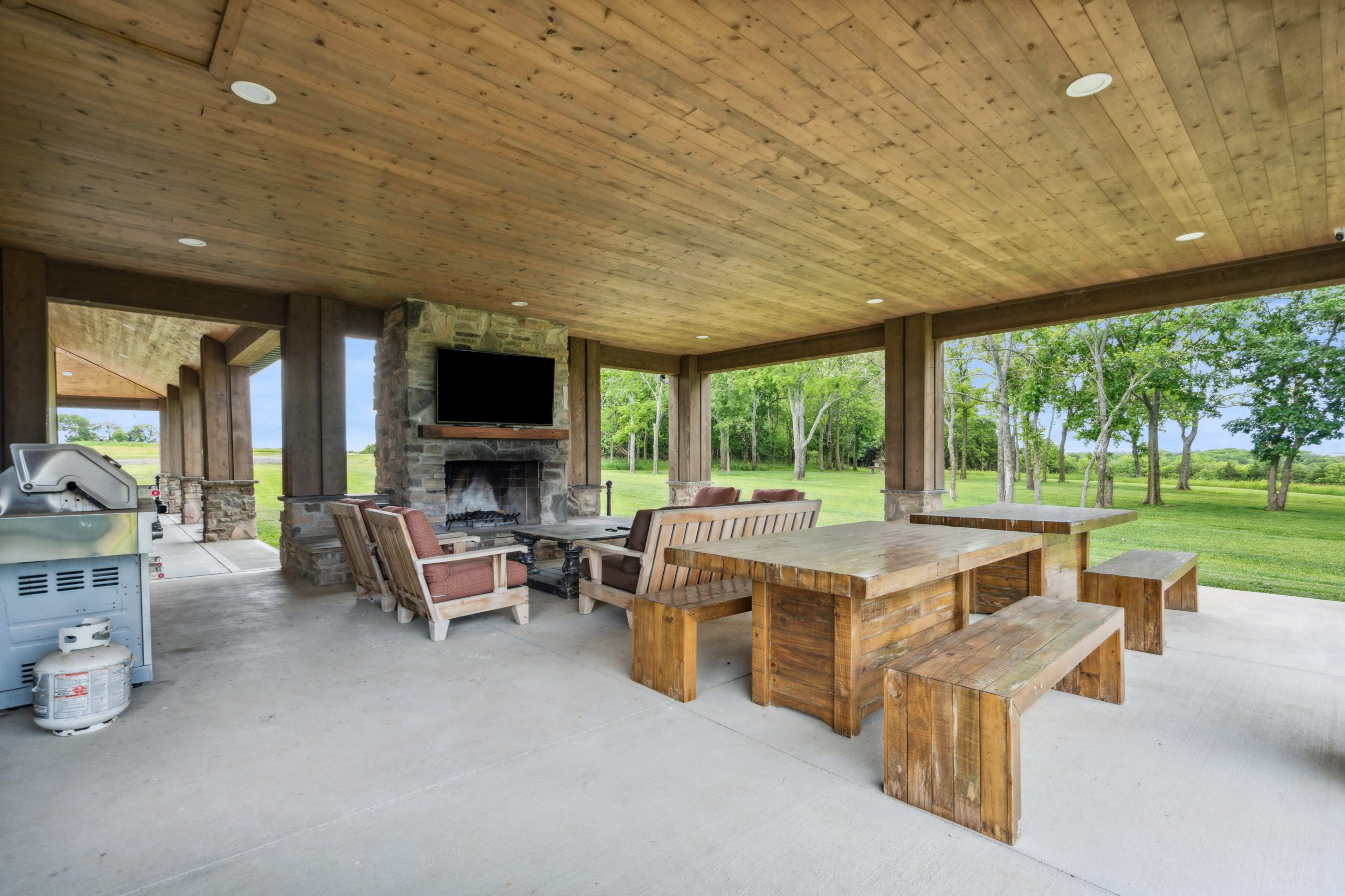 The image shows a covered outdoor area featuring a stone fireplace, wooden seating, a long table, and a grill, with a view of a grassy landscape.