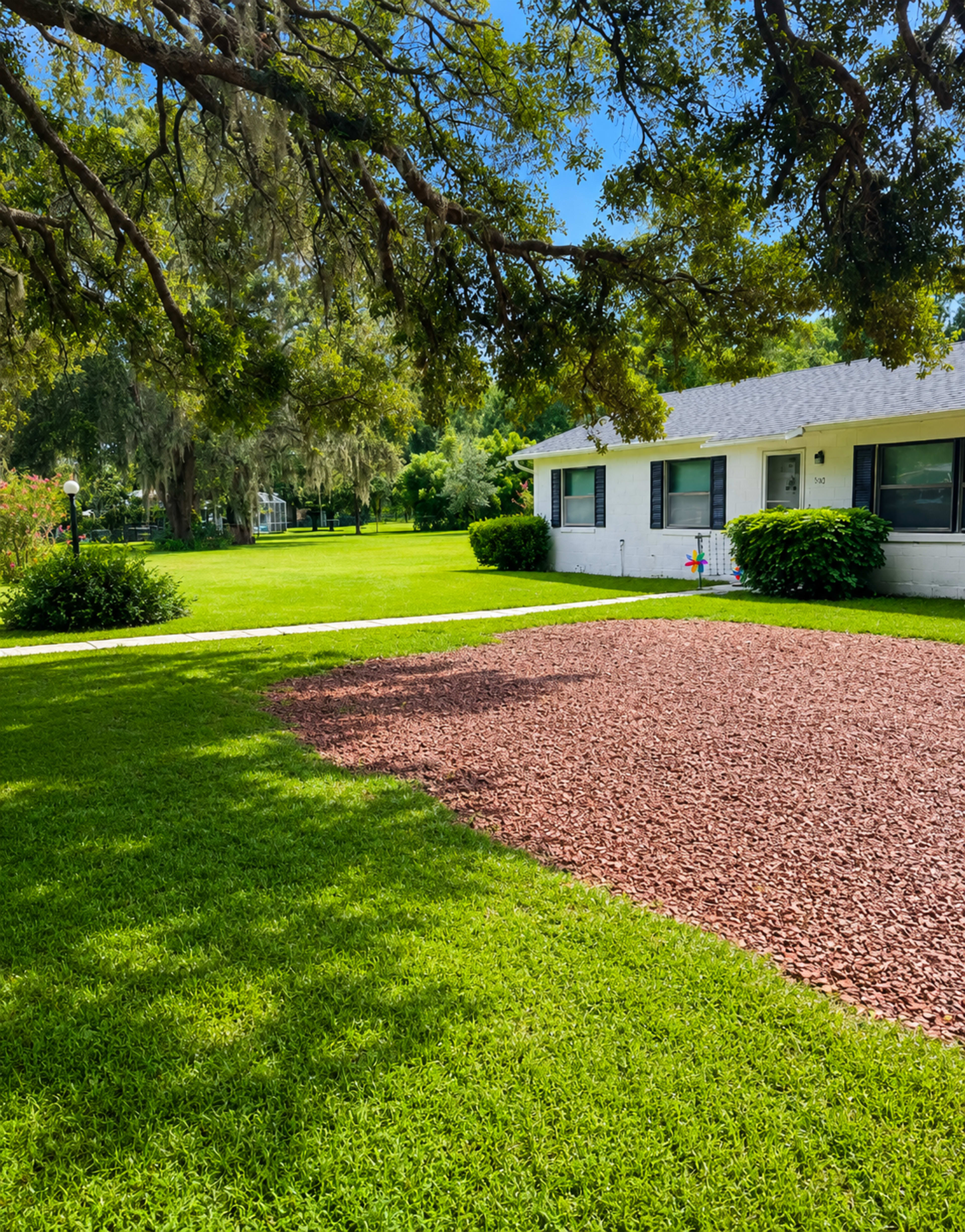 A white house with a gray roof sits beside a gravel pathway on a well-maintained lawn.