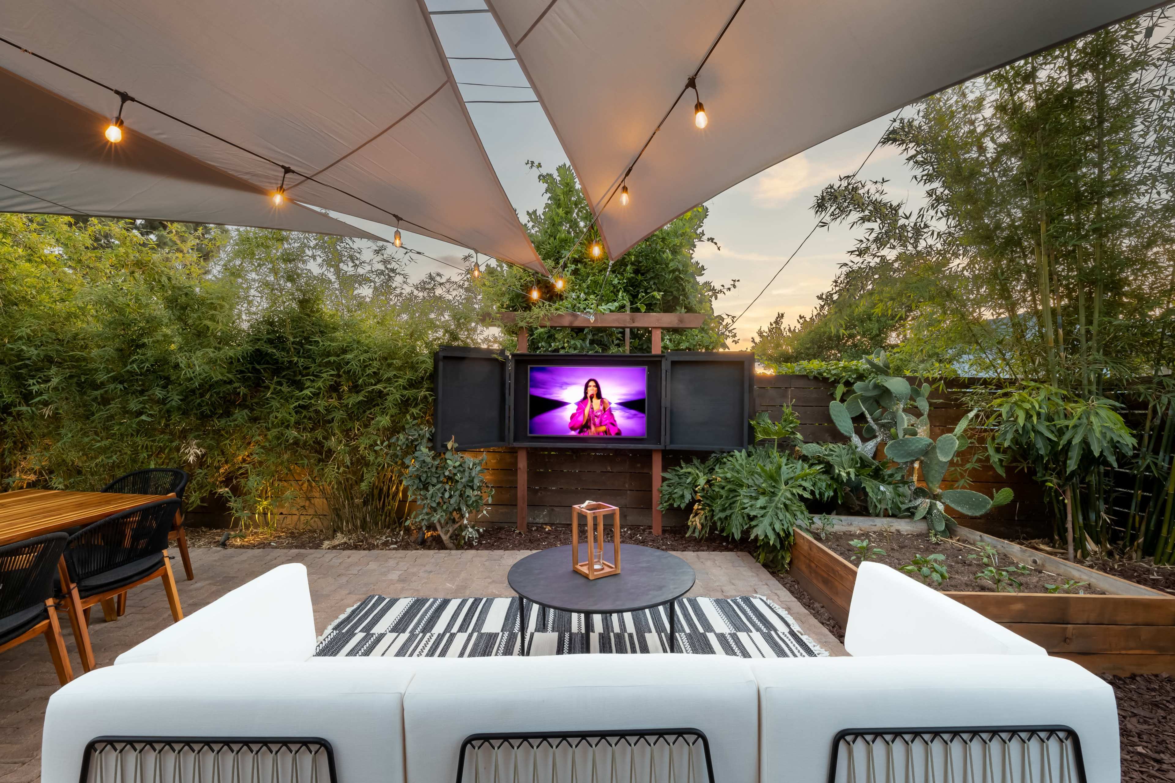 A cozy outdoor patio features a white sofa, a round table, and a television, all under a shaded canopy adorned with string lights and surrounded by greenery.
