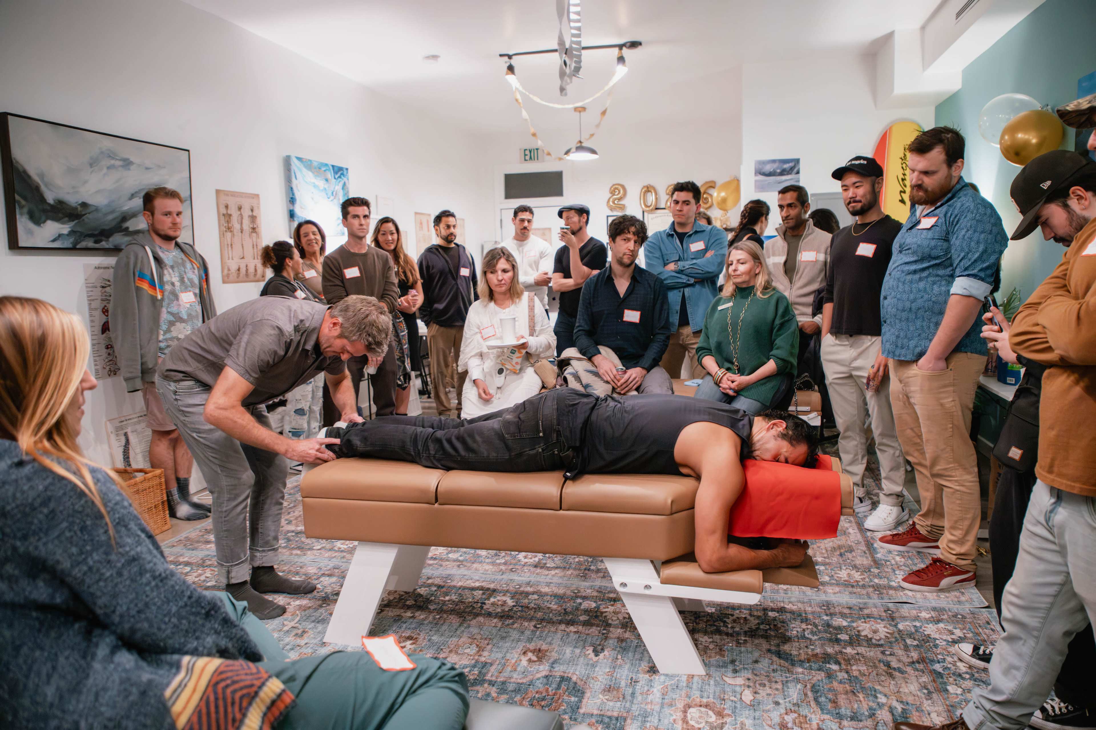 A group of people observes a man receiving a massage on a treatment table in a brightly lit room.