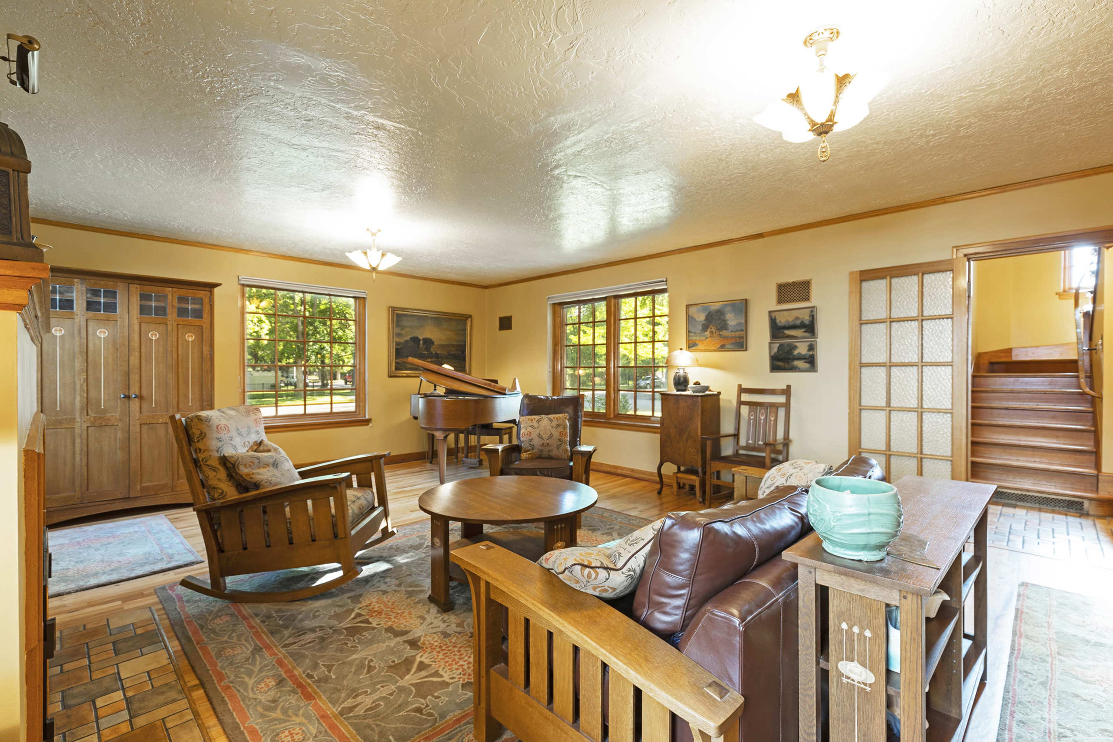 The image shows a cozy living room with wooden furniture, a circular table, and large windows allowing natural light.