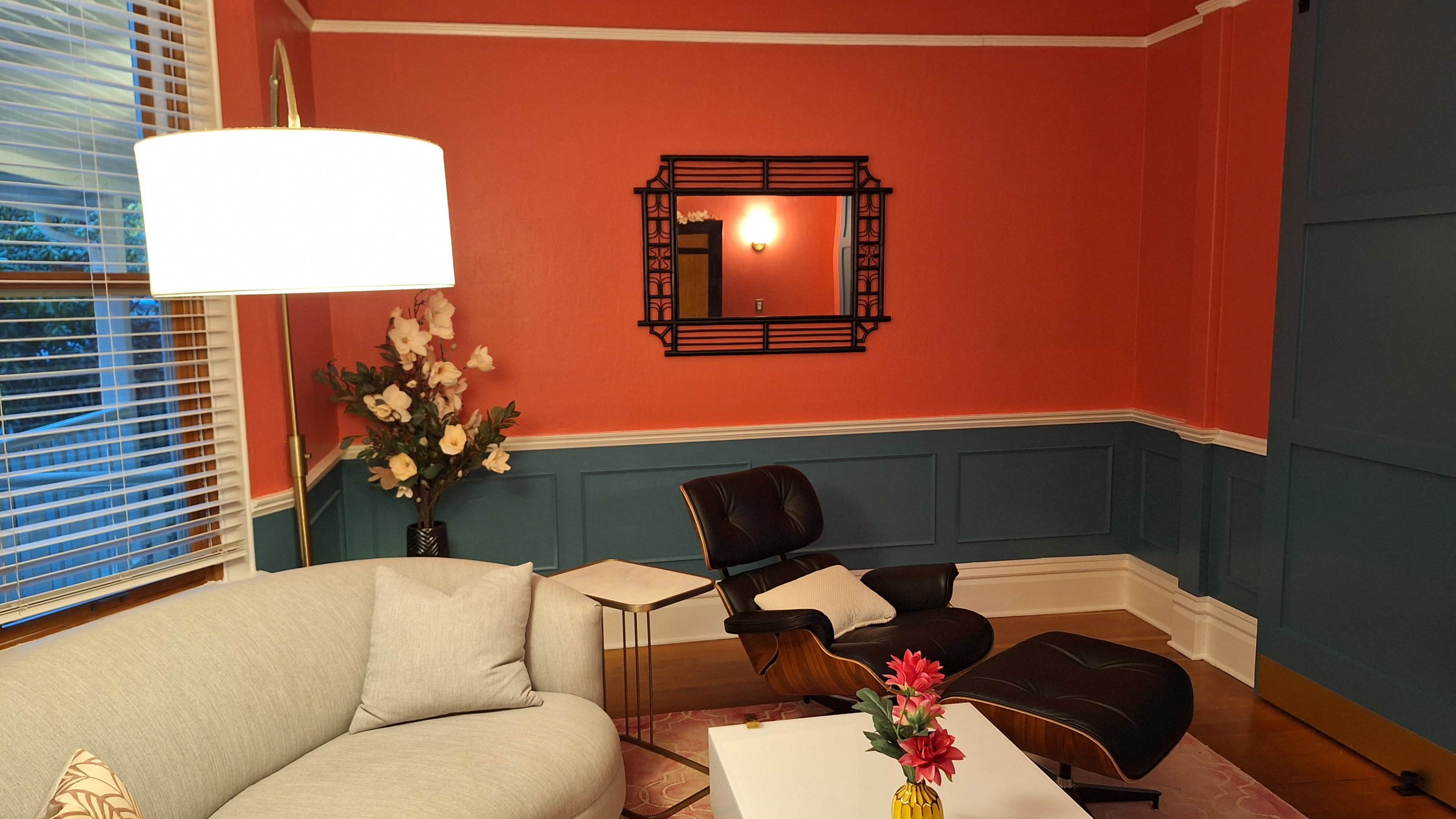The image shows a cozy living room featuring a light-colored sofa, an Eames lounge chair, a small white side table, and a decorative mirror on a brightly colored wall with a floral arrangement nearby.
