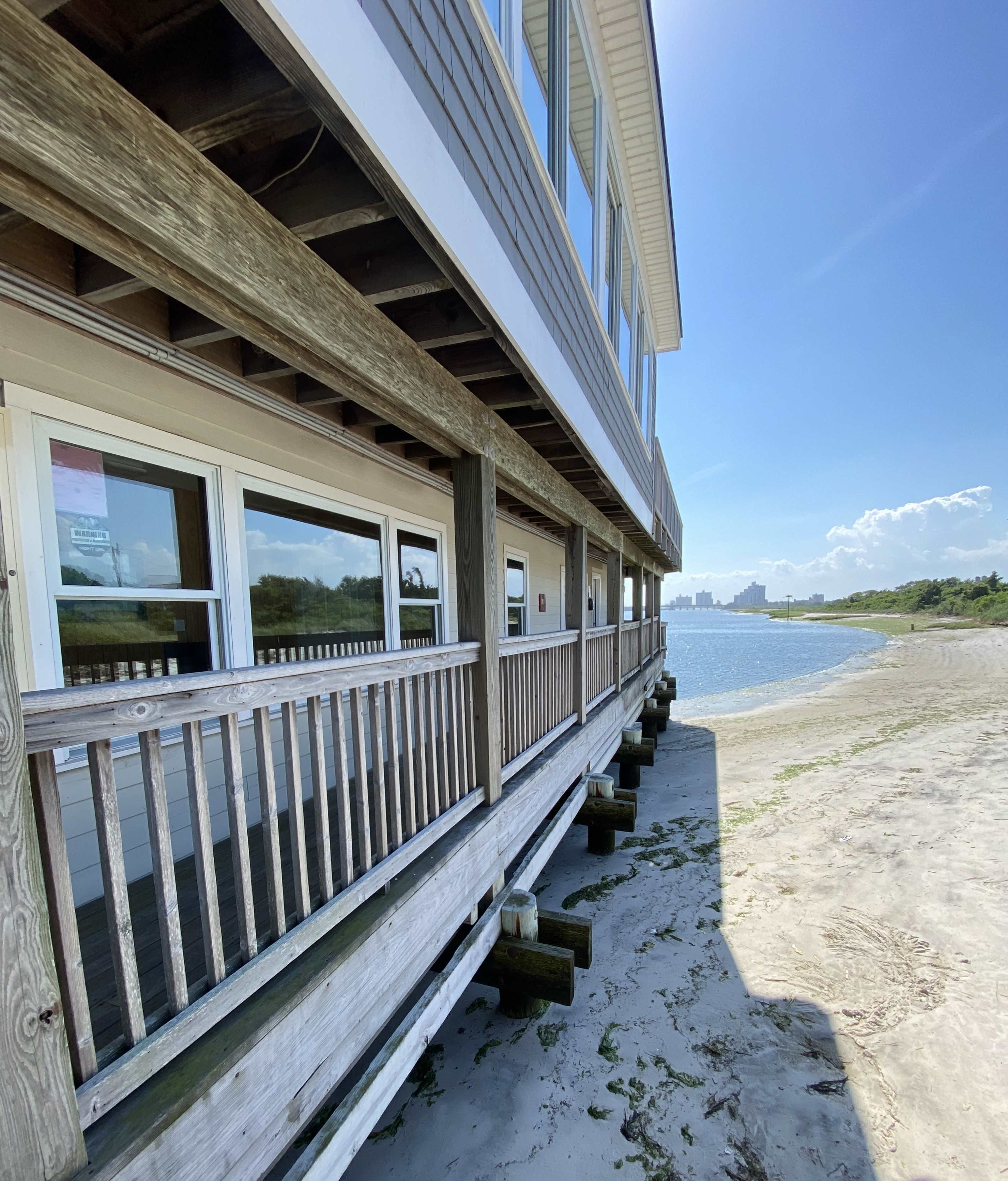 A wooden building sits on stilts along a sandy beach with a view of the water and distant cityscape.