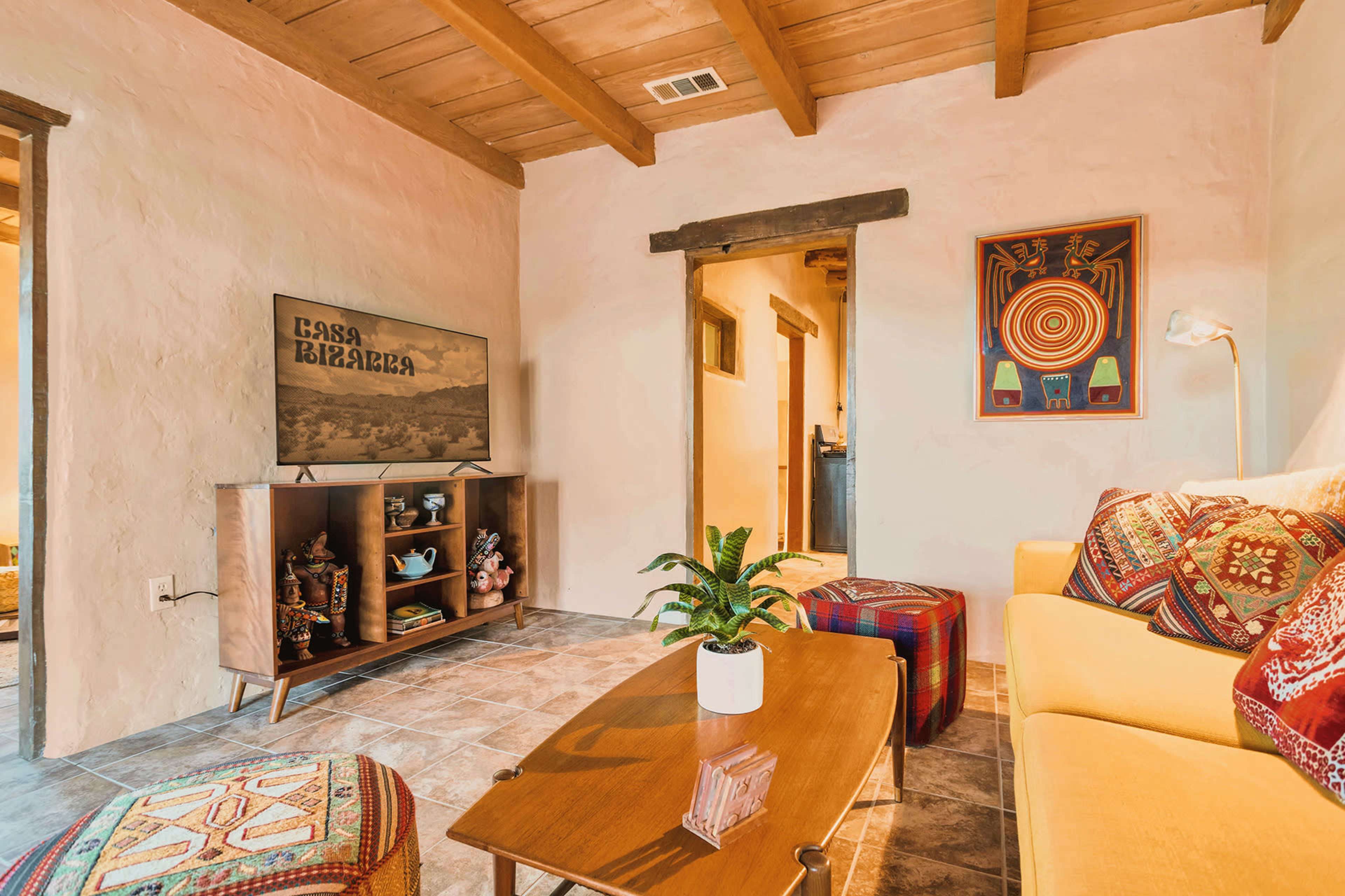 The image shows a cozy living room featuring a yellow sofa, a wooden coffee table, and a television mounted on the wall with decorative art and plants.