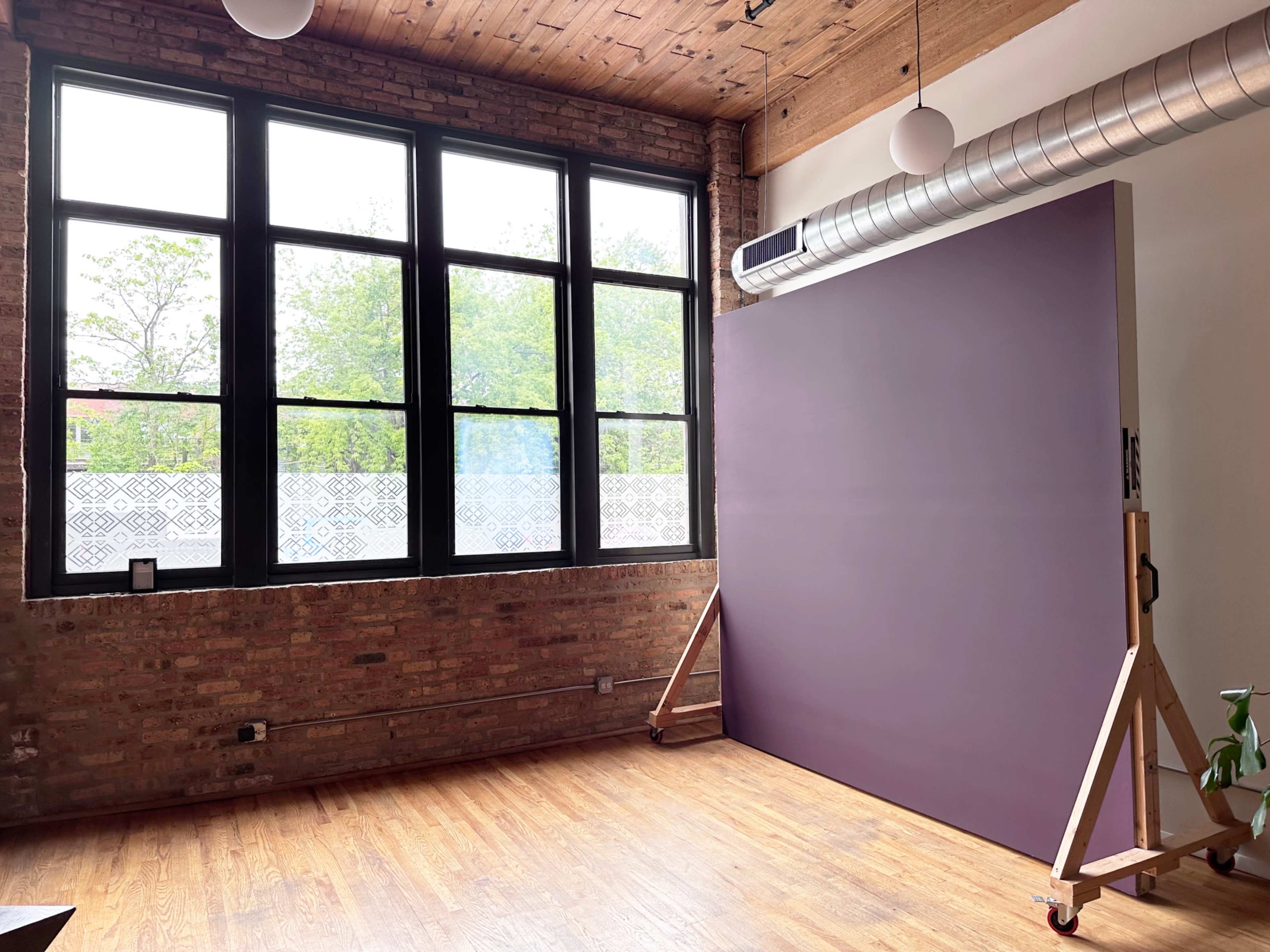The image shows a room with exposed brick walls, large windows allowing natural light, and a purple backdrop on wheels.
