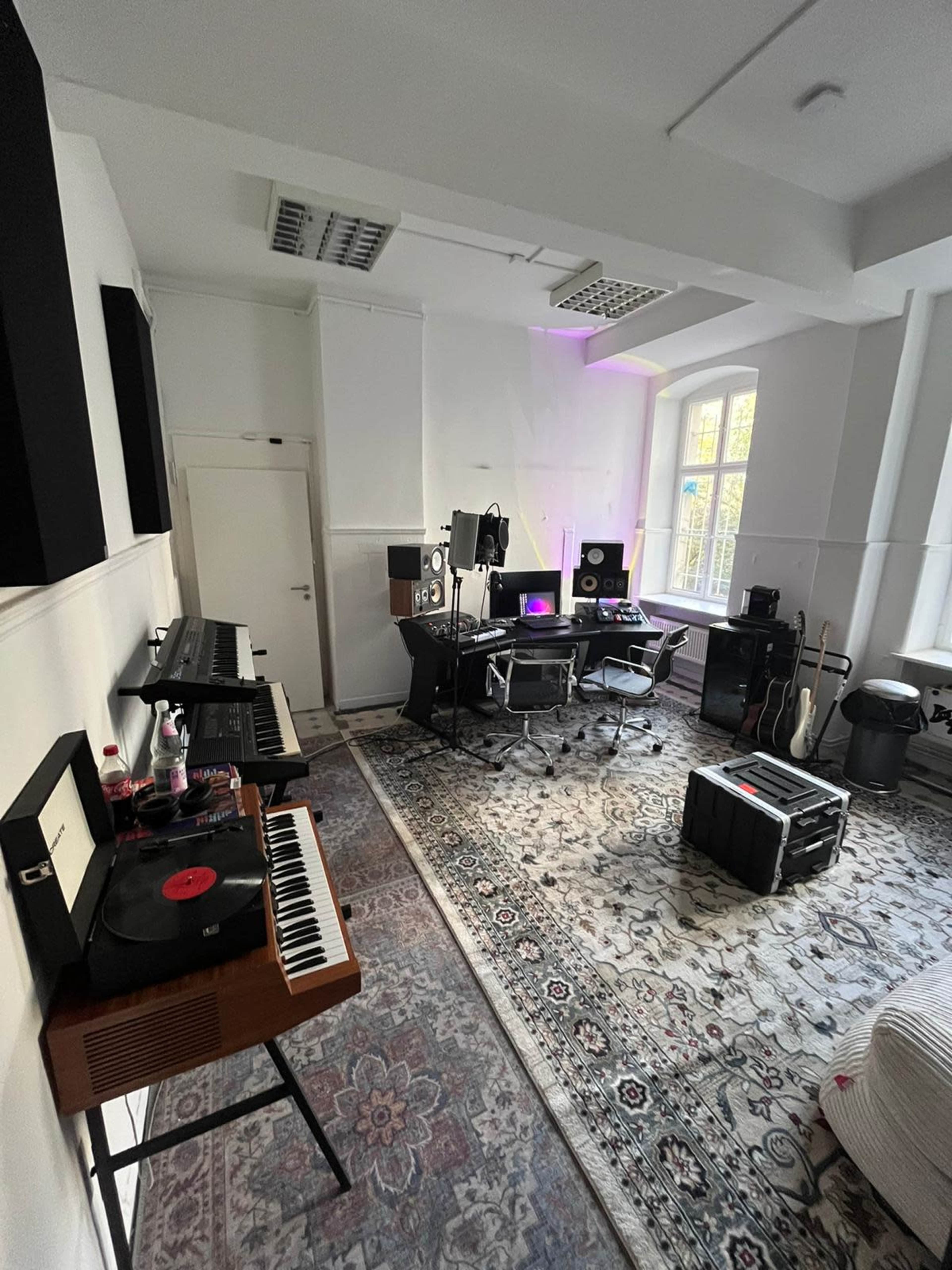 The image shows a music production studio featuring keyboards, recording equipment, a desk with monitors, and a large area rug.