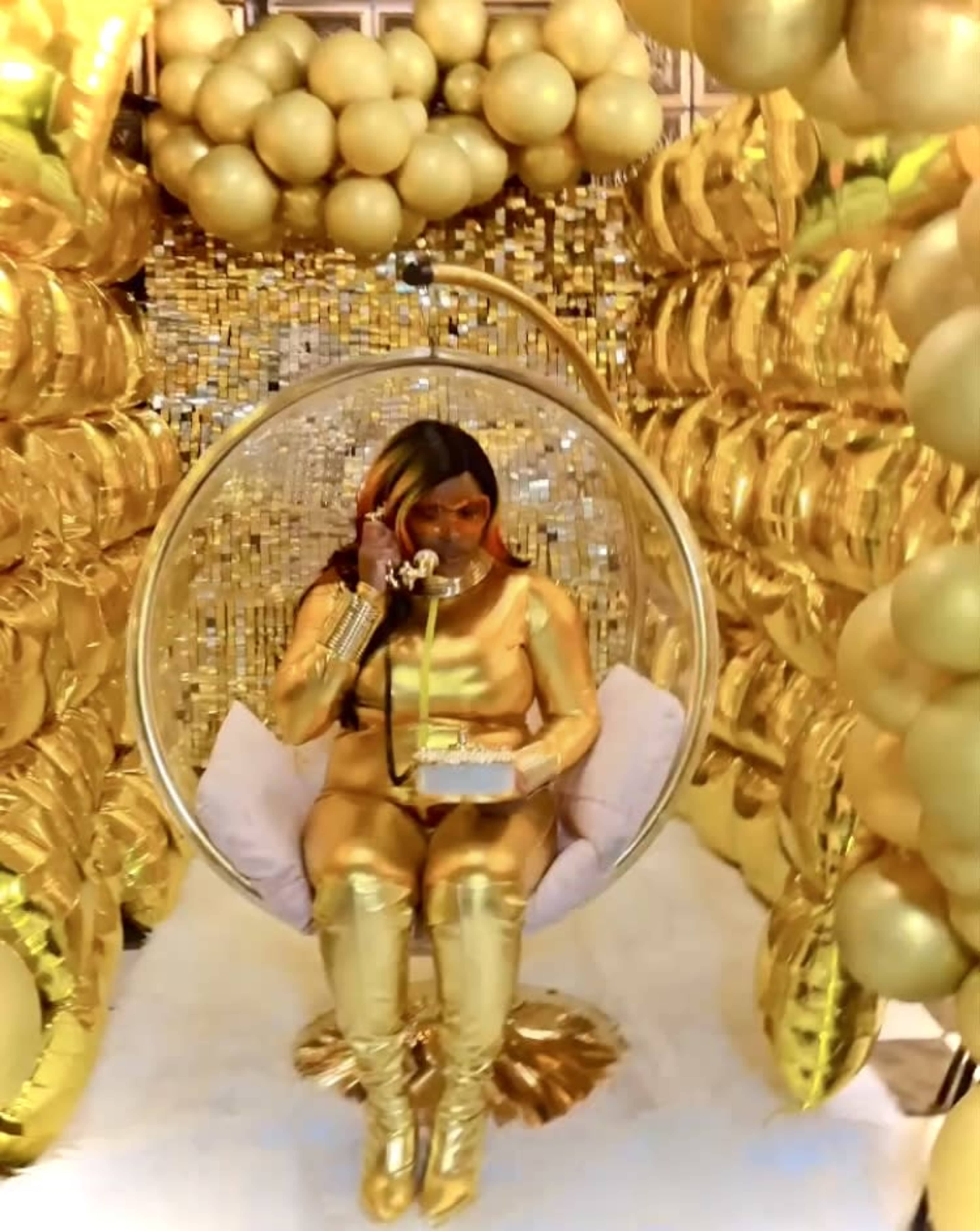 A person dressed in gold sits in a transparent spherical chair surrounded by golden balloons and shiny decor.