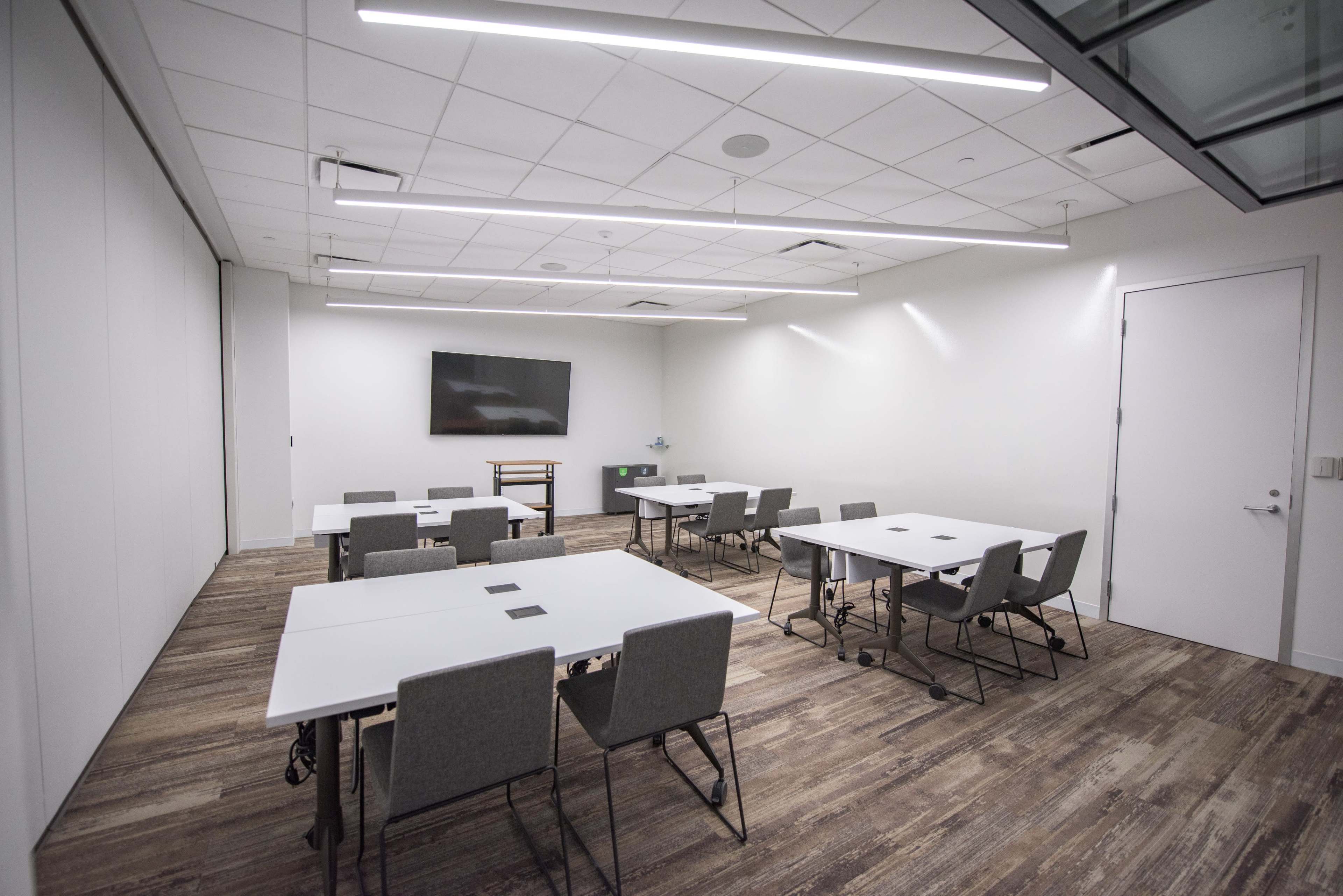 A modern meeting room features multiple tables and chairs arranged around a central space, with a wall-mounted screen and soft lighting.