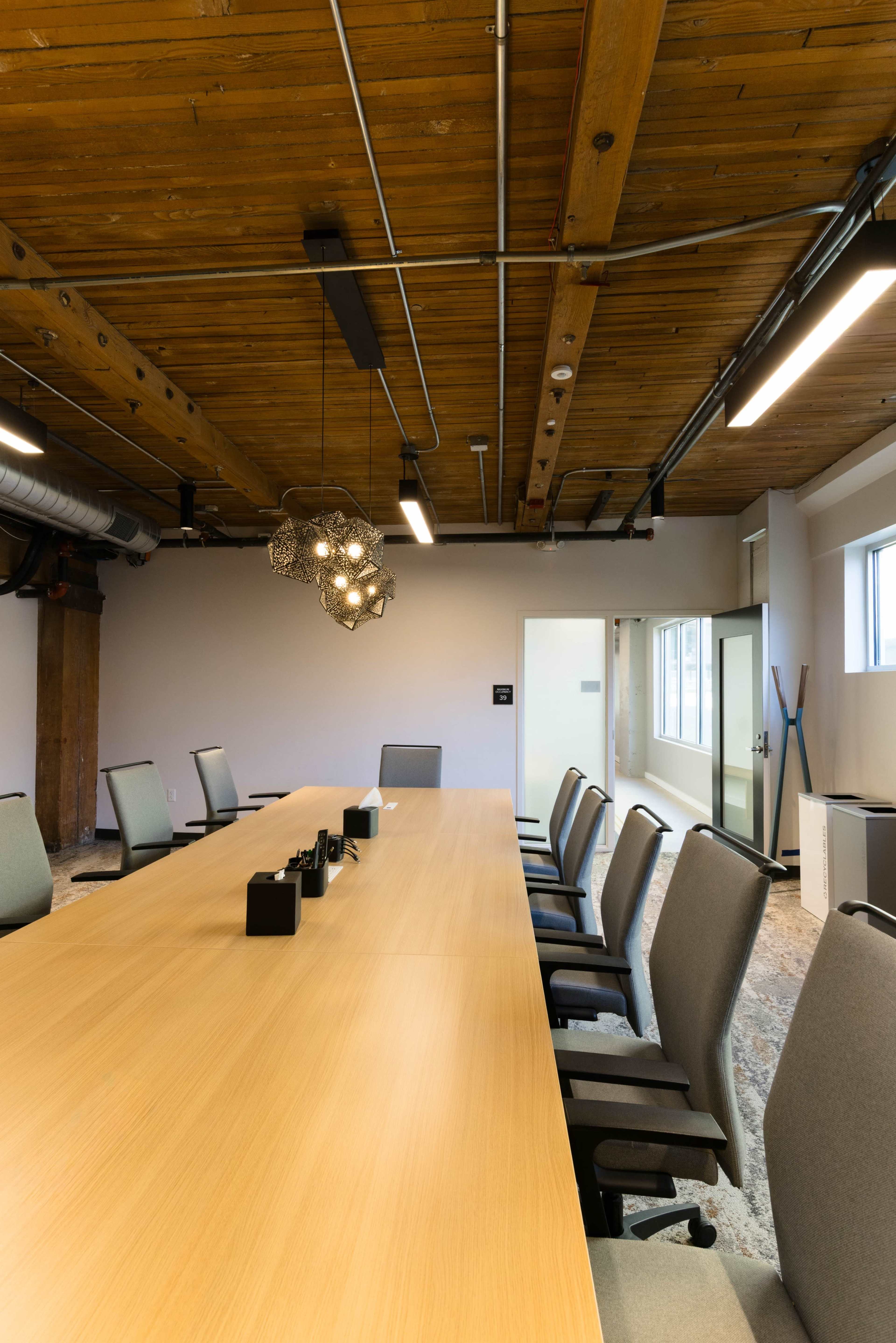 A modern conference room features a long wooden table surrounded by ergonomic chairs and is illuminated by a unique pendant light.