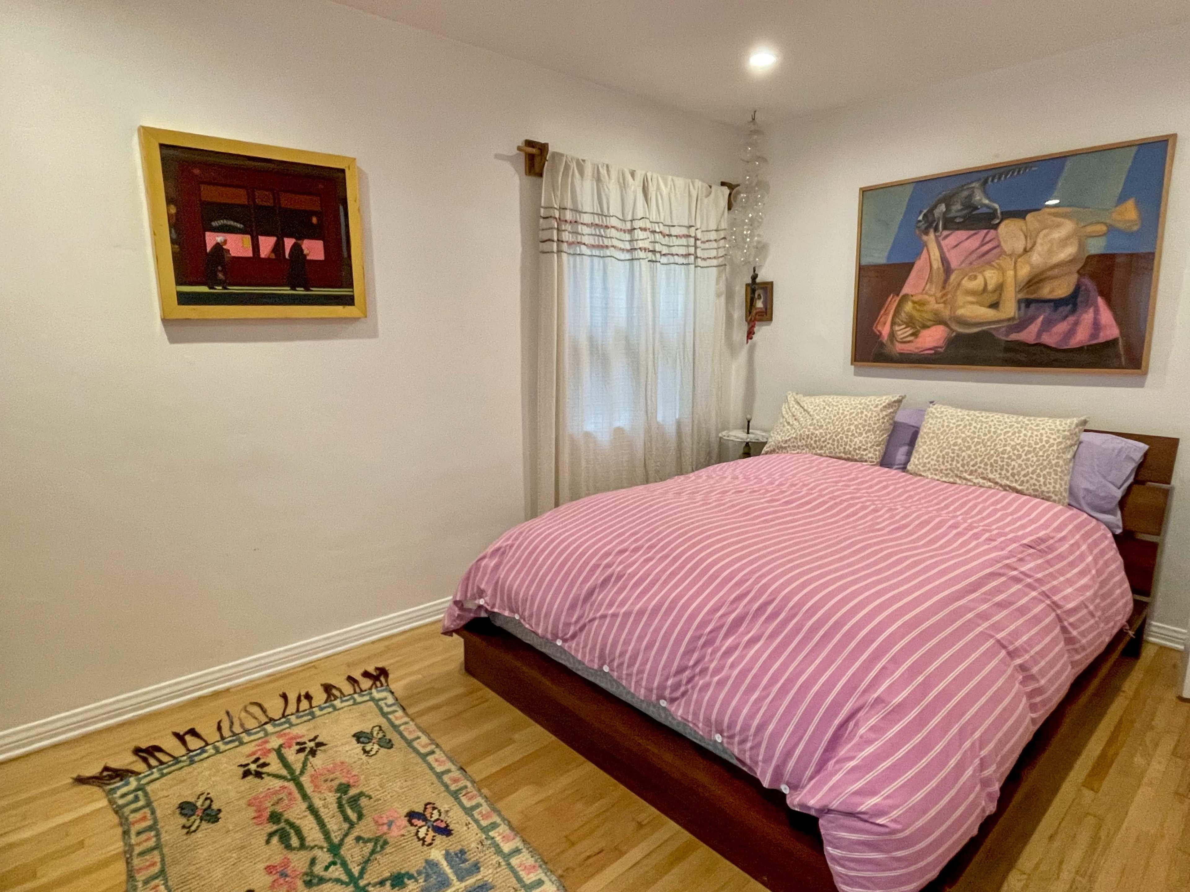 A bedroom with a wooden bed covered in a pink striped duvet, decorated with two patterned pillows, and featuring artwork and a rug on the wooden floor.