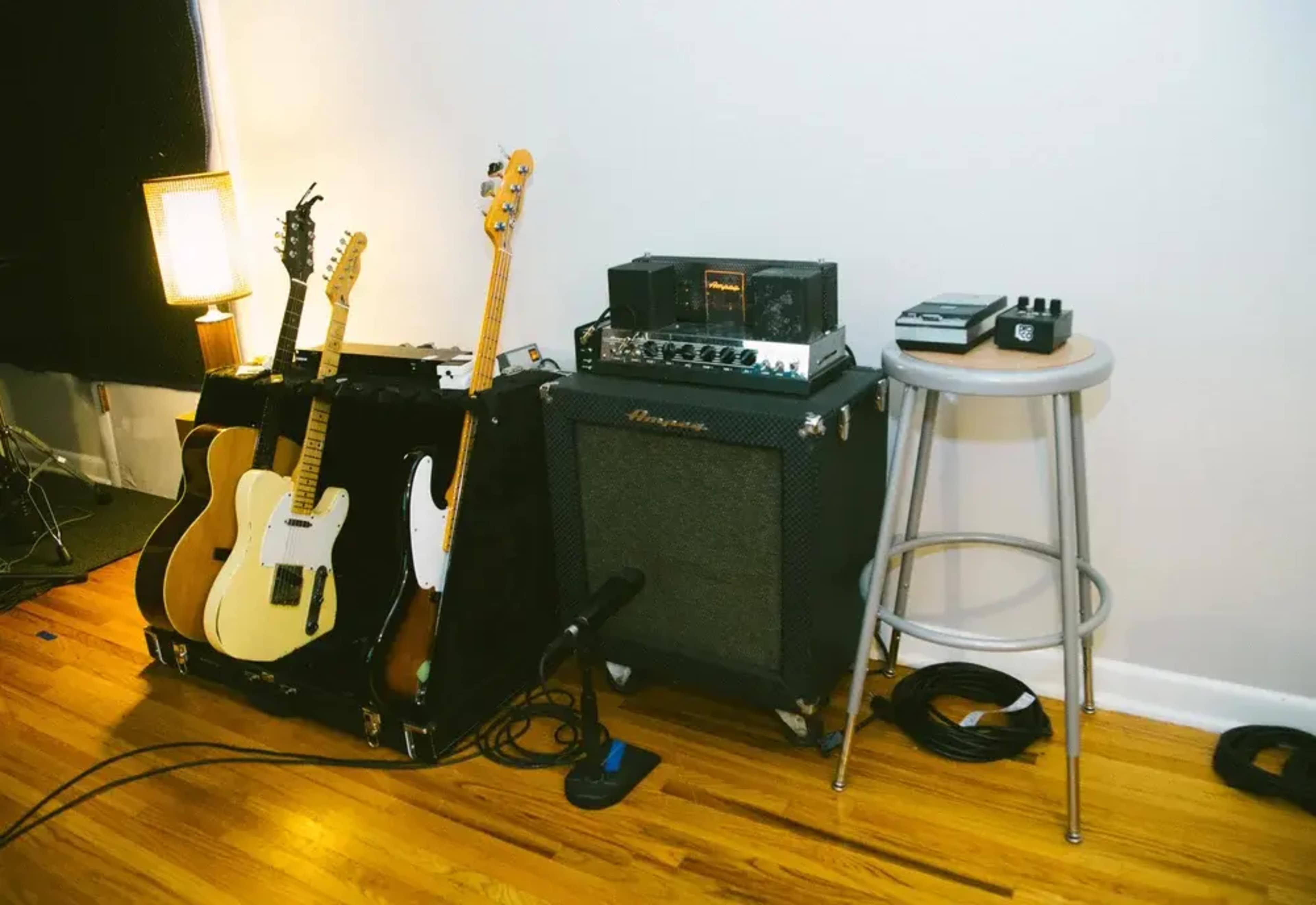 The image shows two electric guitars leaning against a case next to an amplifier and a stool, with cables lying on the wooden floor.
