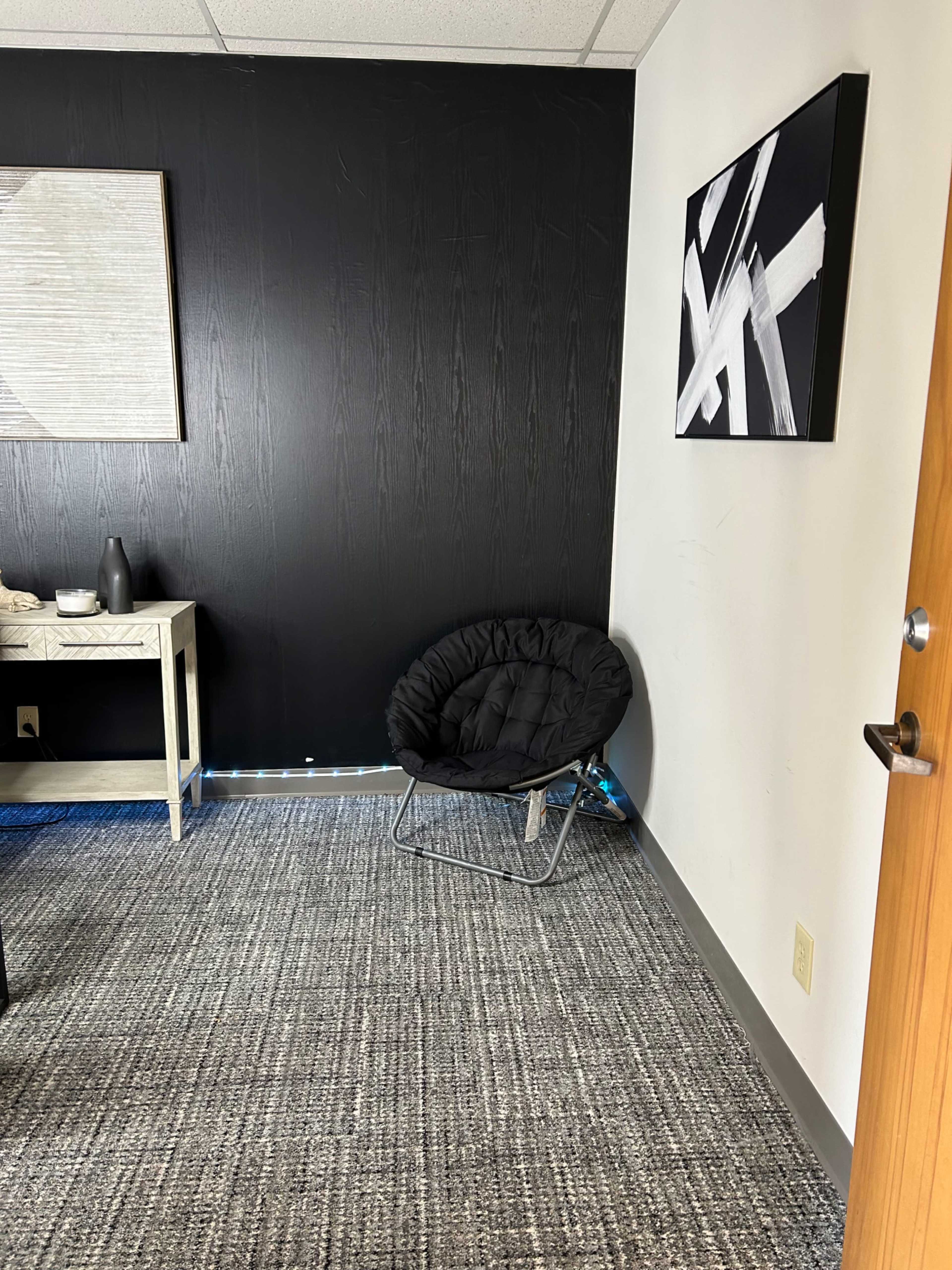 A small, modern corner of a room featuring a black fabric chair, a side table with decor, and abstract artwork on the wall.