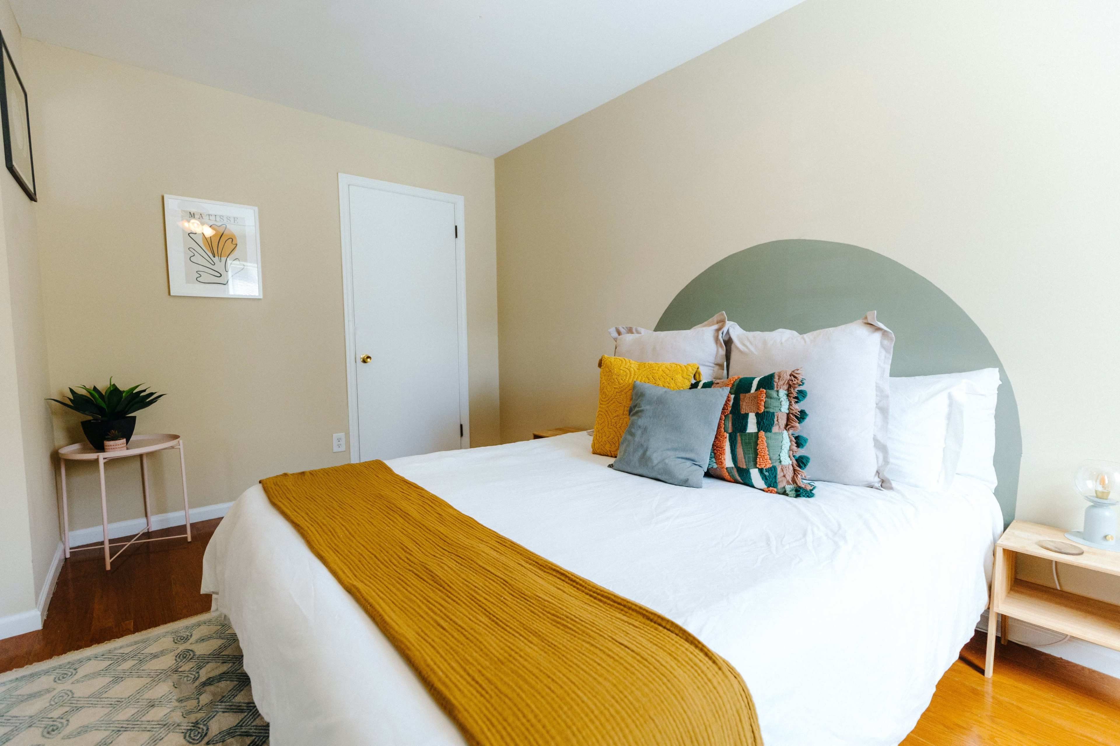 A brightly lit bedroom features a bed with white linens and colorful pillows, a small nightstand, and a potted plant.
