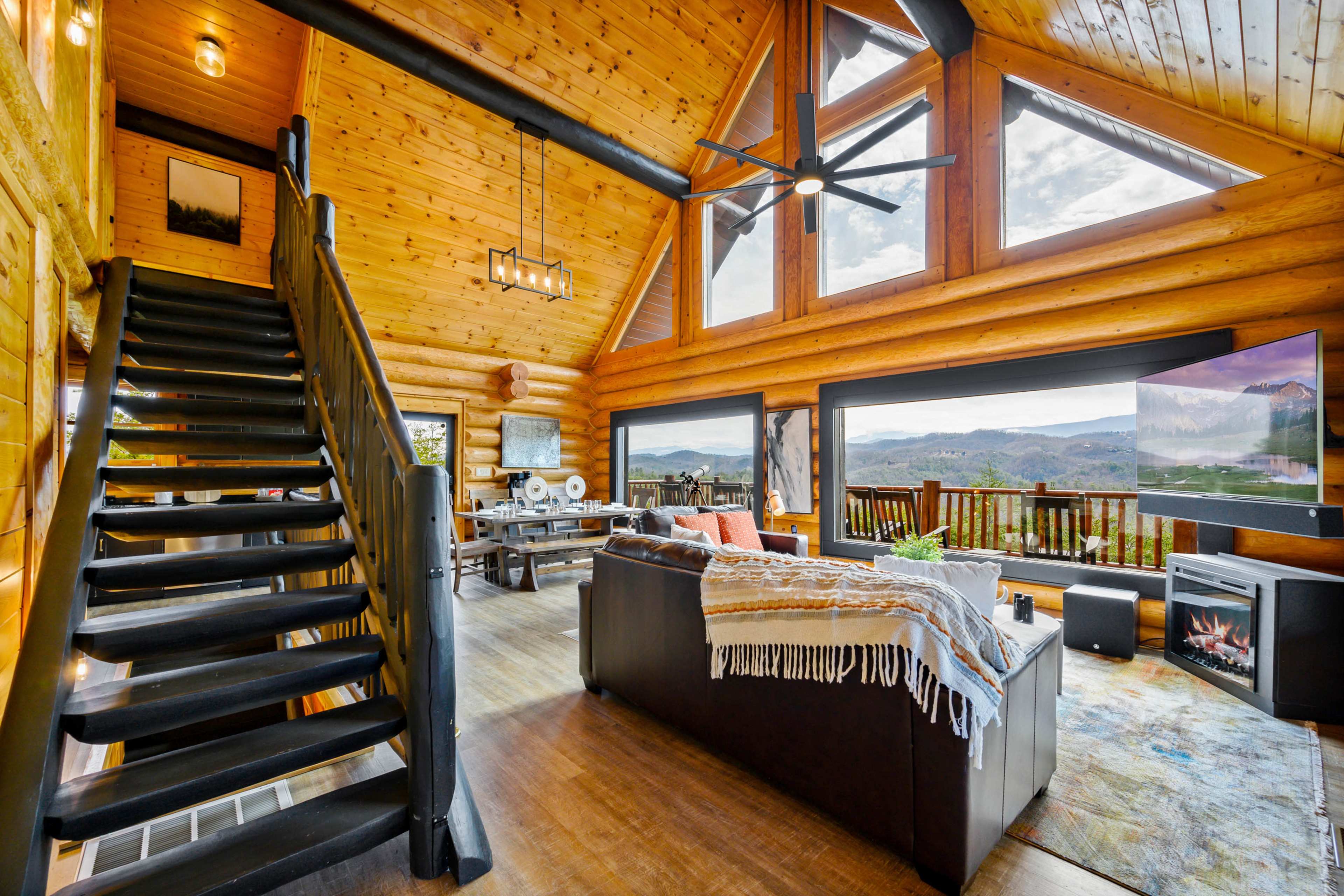The interior of a log cabin features a spacious living area with large windows, a comfortable couch, a staircase leading to an upper level, and a television with a fireplace nearby.