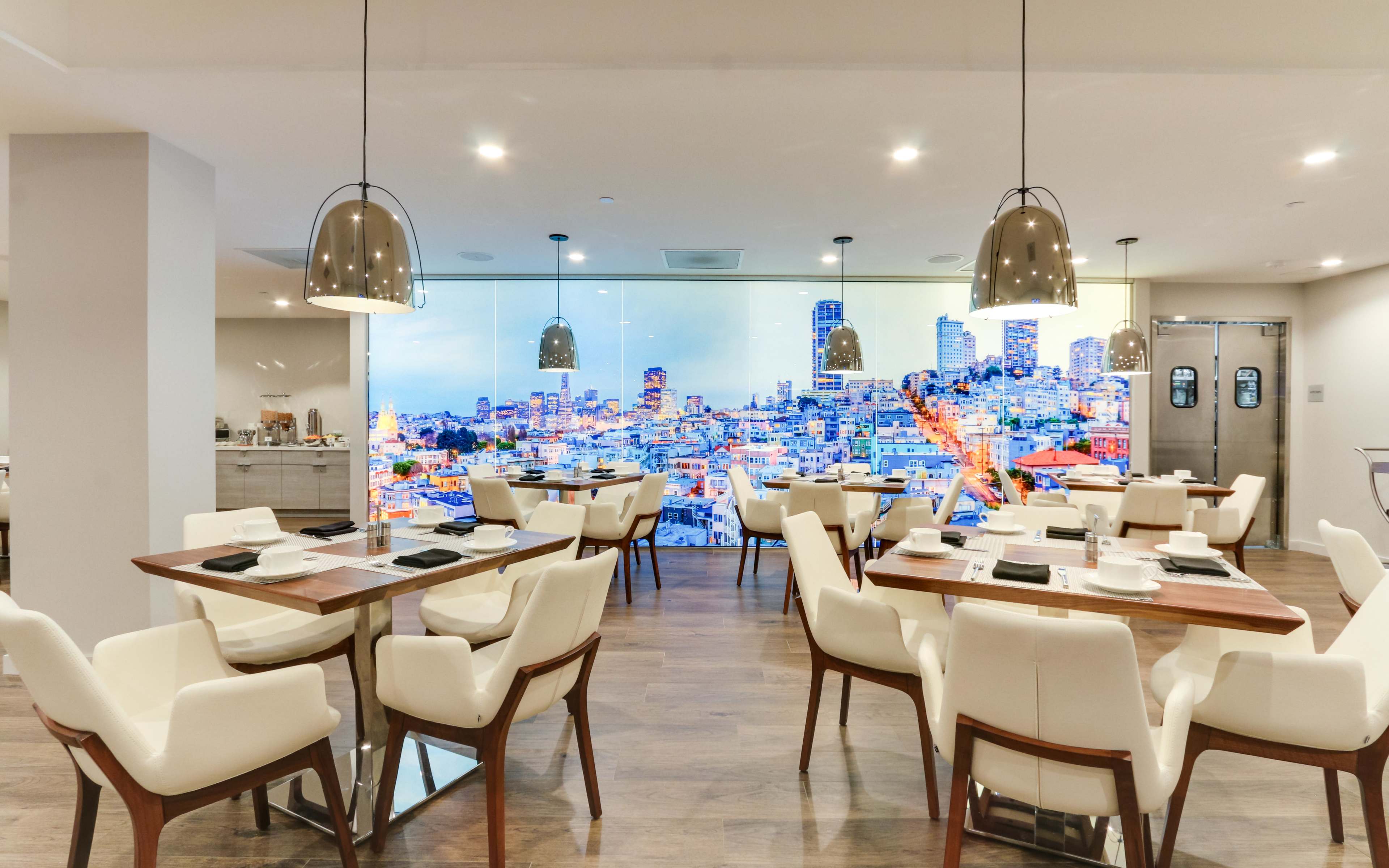 The image shows a modern dining area with white chairs and wooden tables, featuring a large mural of a city skyline on the wall.