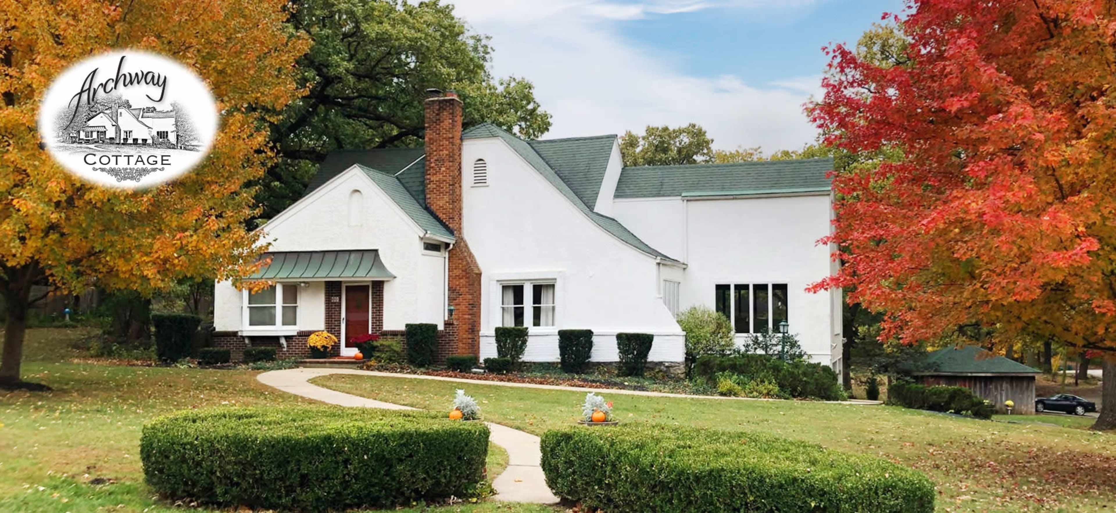 A white cottage with a green roof and brick chimney is surrounded by a well-manicured lawn and colorful autumn trees.