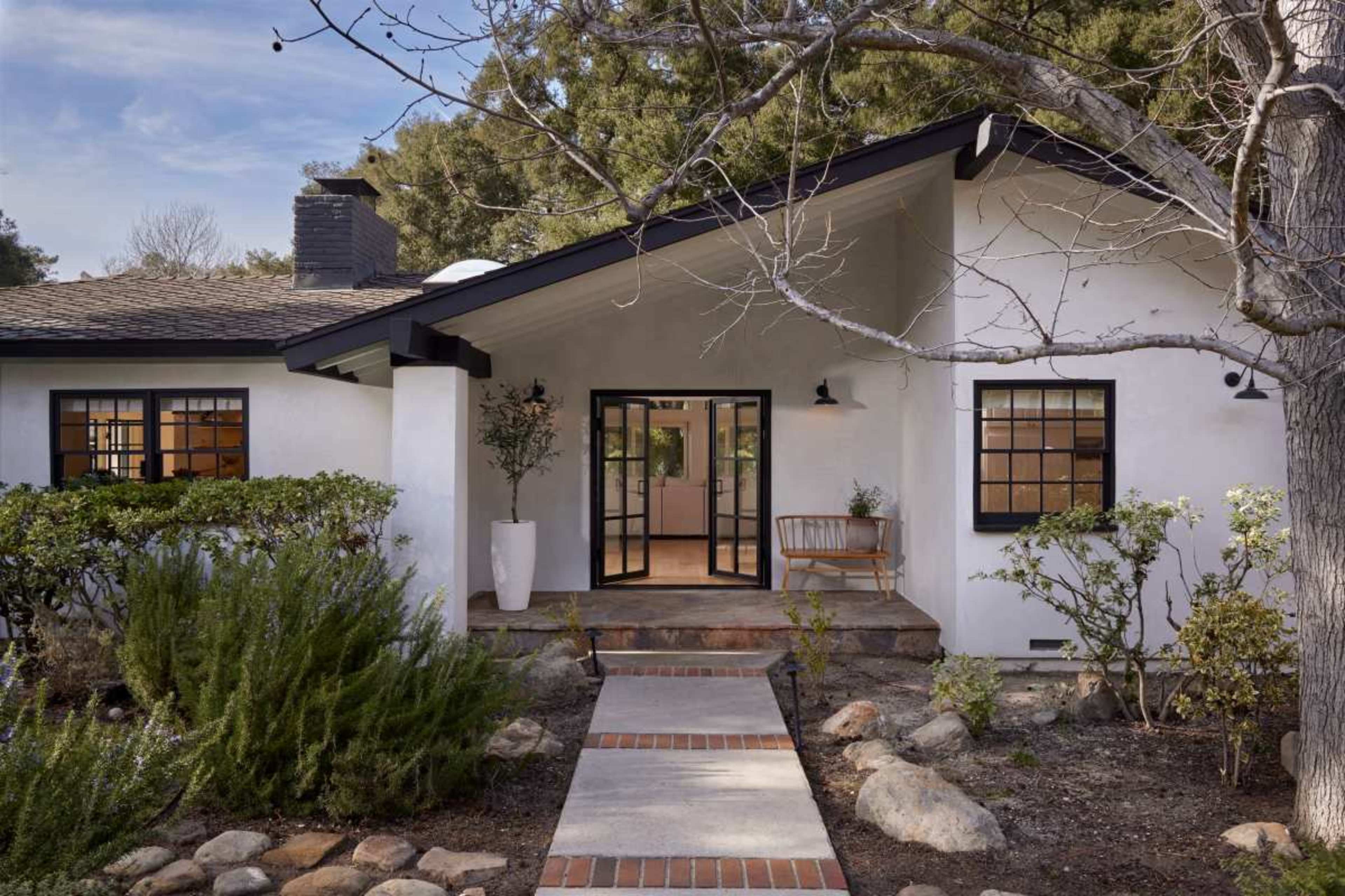 A landscaped pathway leads to the front door of a modern, single-story house with a black roof and white exterior.