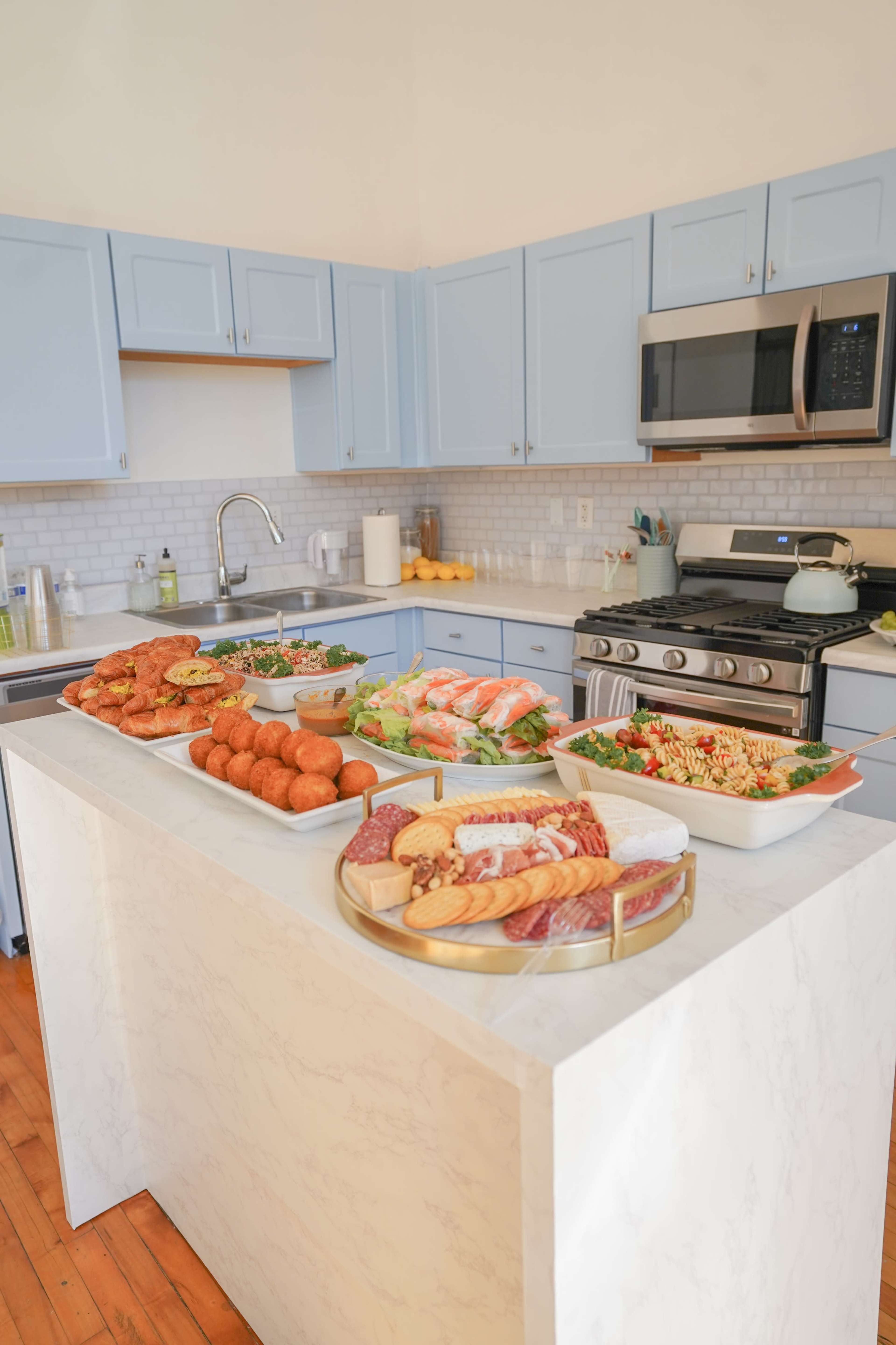 A kitchen island is filled with a variety of prepared foods and dishes, including salads, meats, and pastries, in a brightly lit kitchen with blue cabinetry.