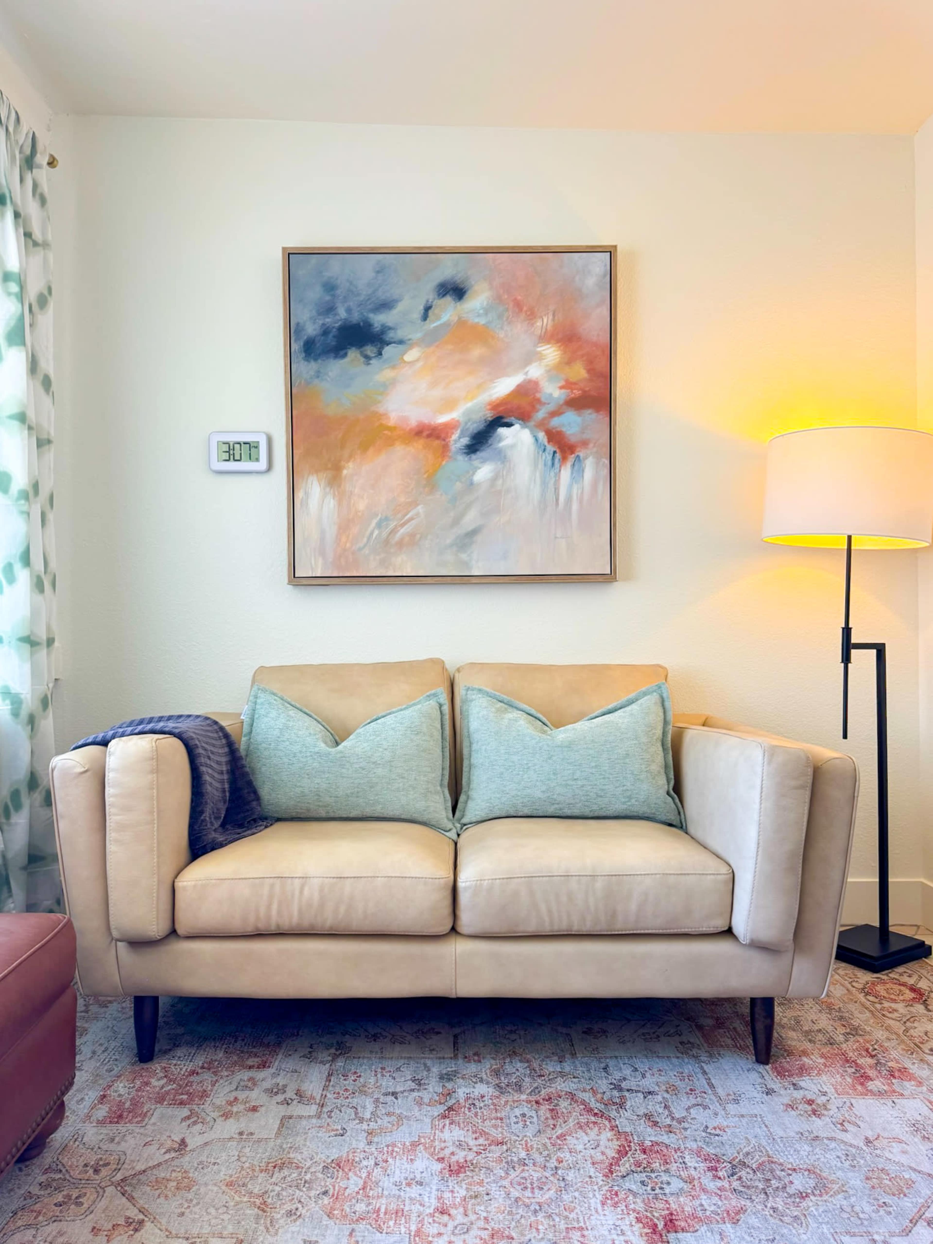 A light-colored sofa with two turquoise cushions is positioned against a wall featuring an abstract painting and a floor lamp.