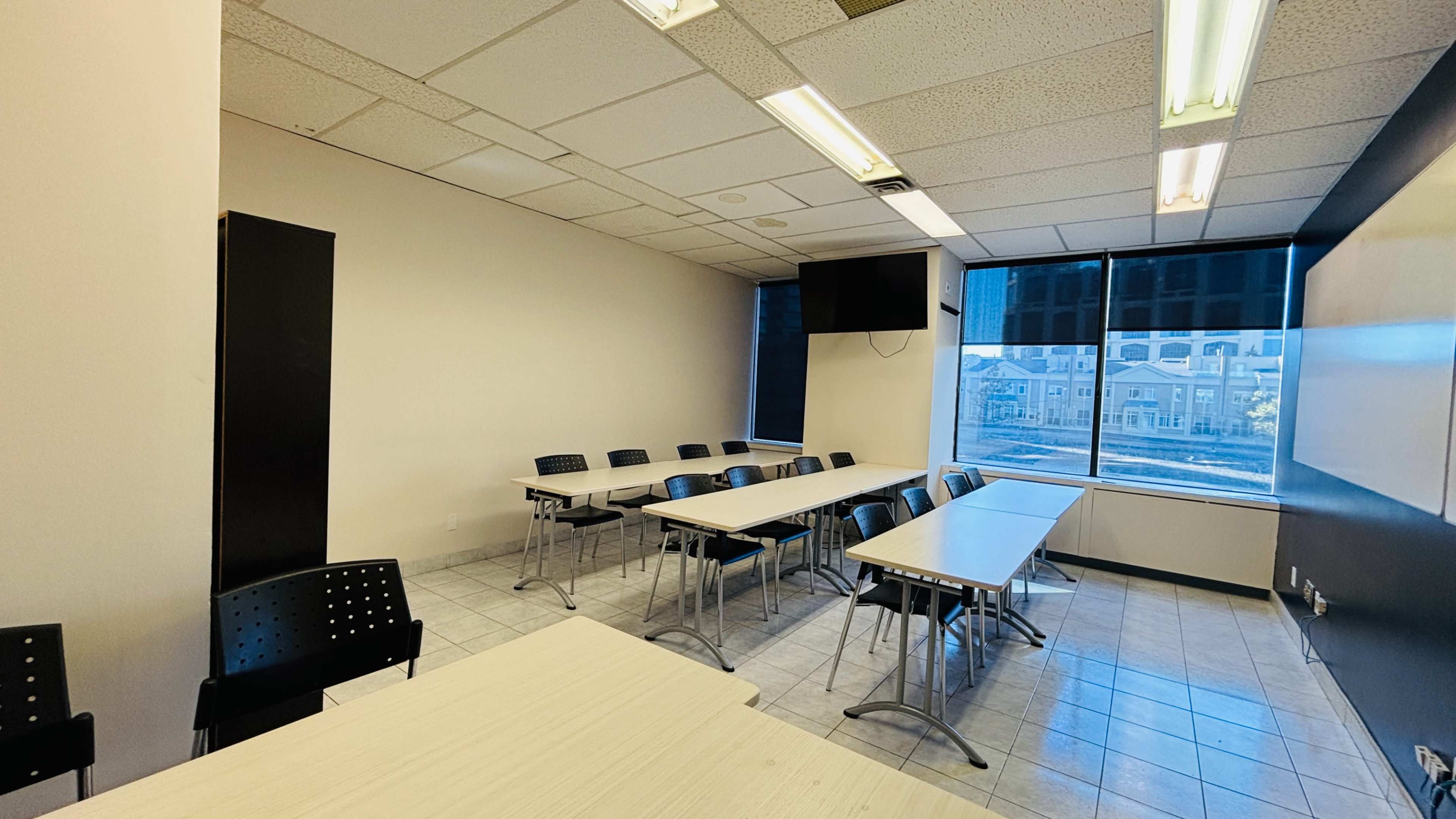 The image shows a classroom with multiple tables arranged in a U-shape, black chairs, and a large window providing natural light.