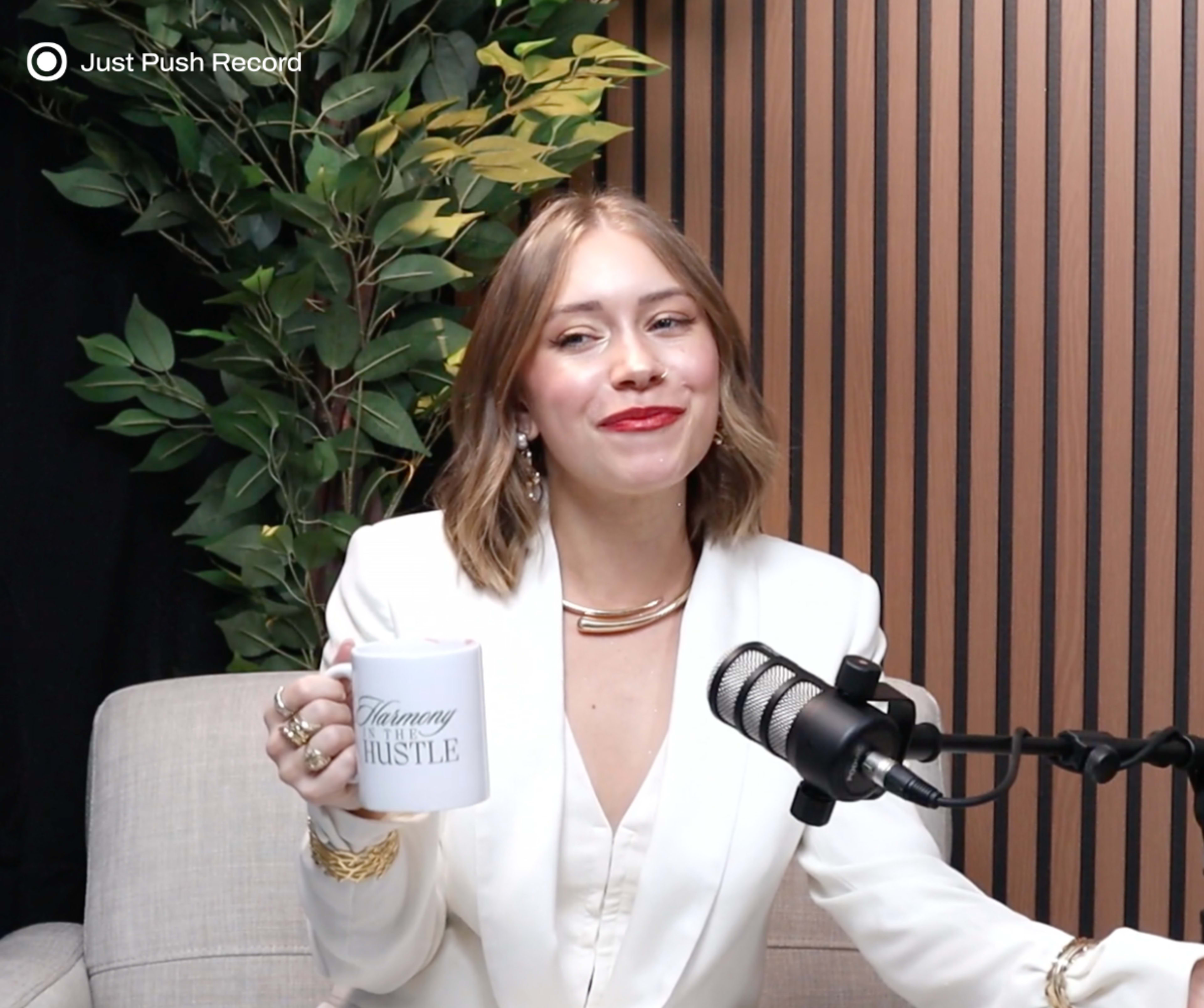 A woman in a white blazer holds a coffee mug while sitting in front of a microphone against a backdrop of green plants and wooden paneling.
