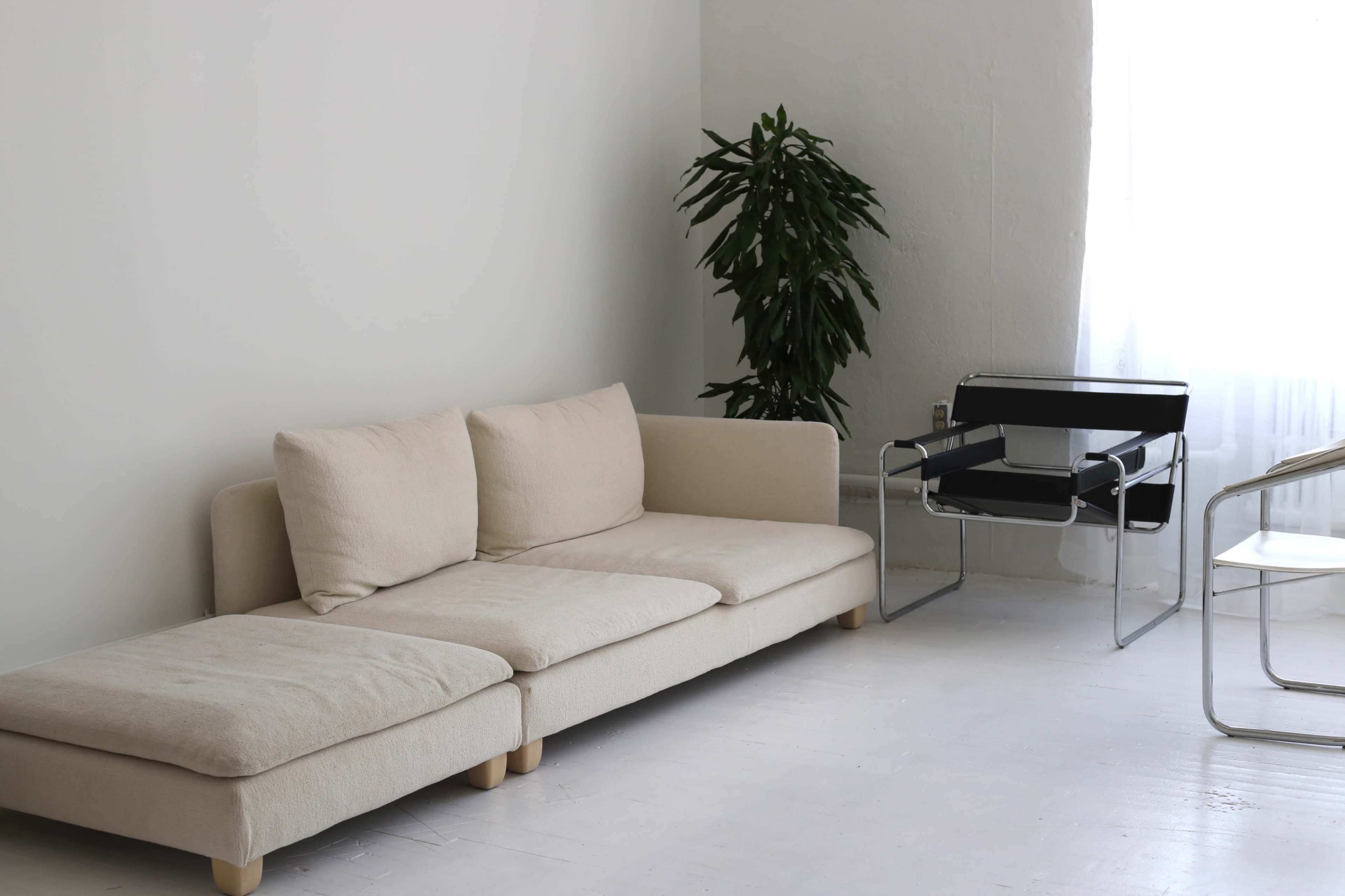 The image depicts a minimalist living space featuring a beige couch, a black chair with a chrome frame, and a plant in the corner.