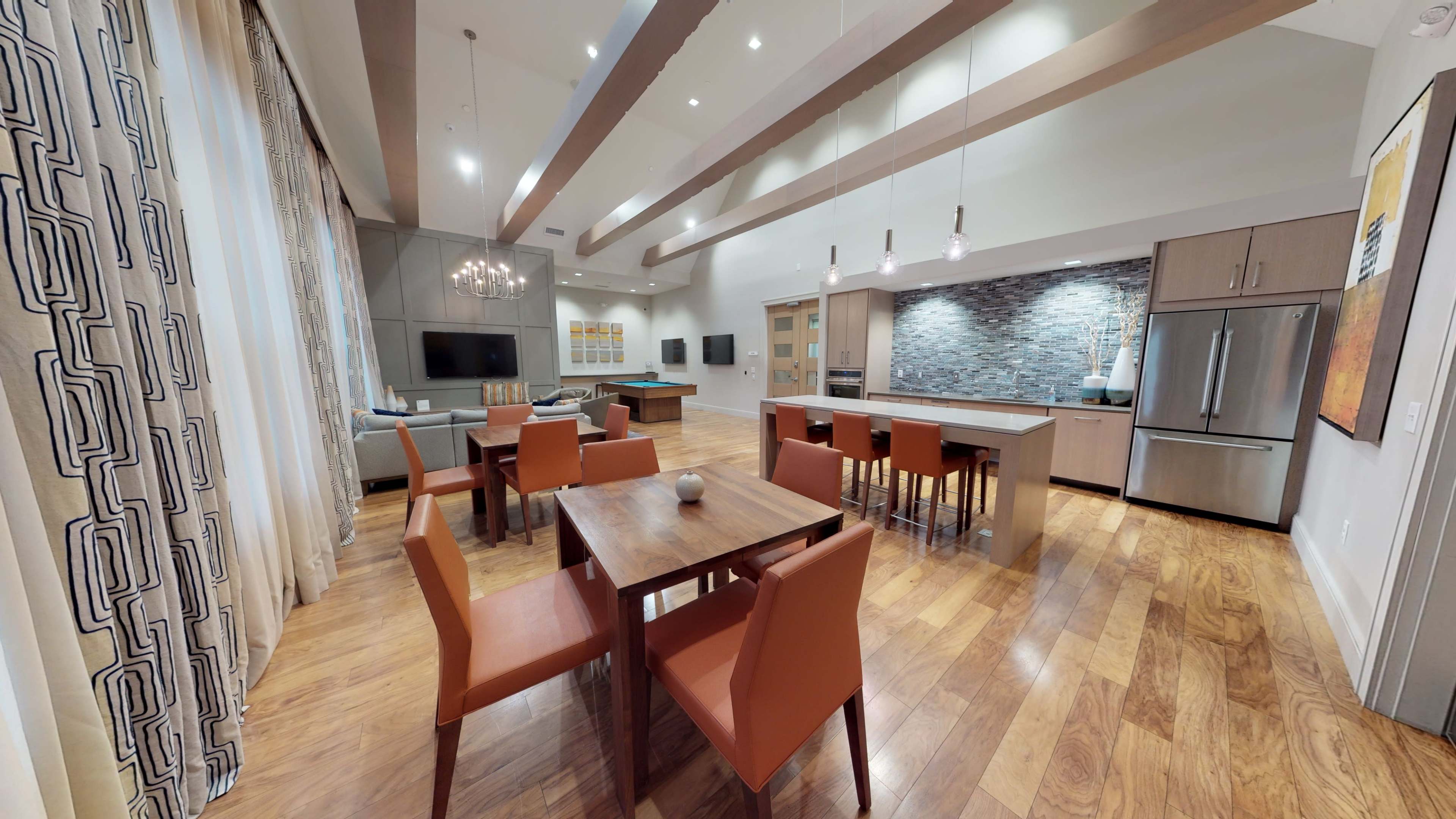 A spacious, modern kitchen and dining area with a wooden dining table surrounded by orange chairs and a kitchen equipped with stainless steel appliances and a stone backsplash.