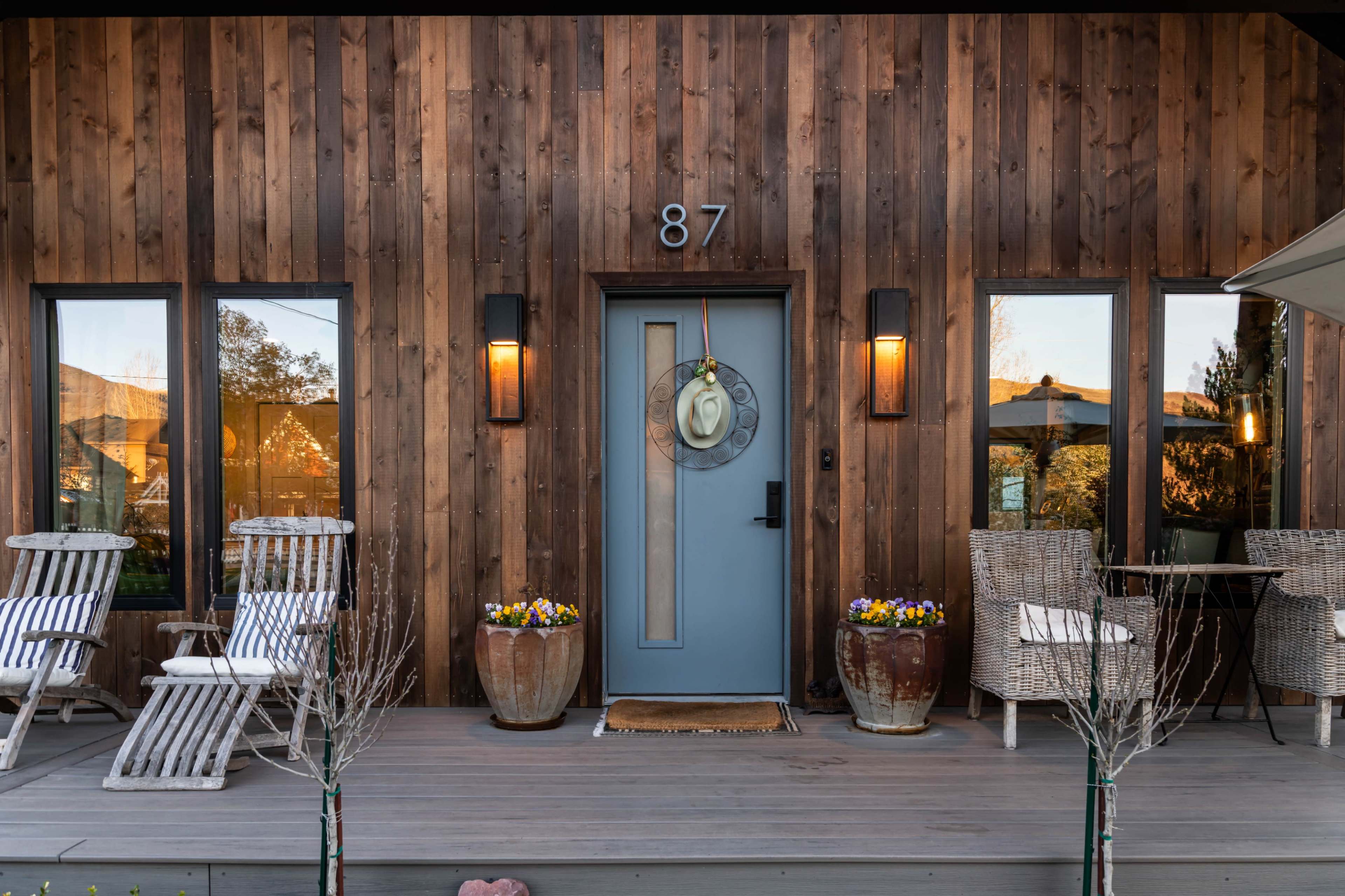 The image shows a wooden house facade with a blue front door, number 87, flanked by outdoor seating and decorative pots on a porch.