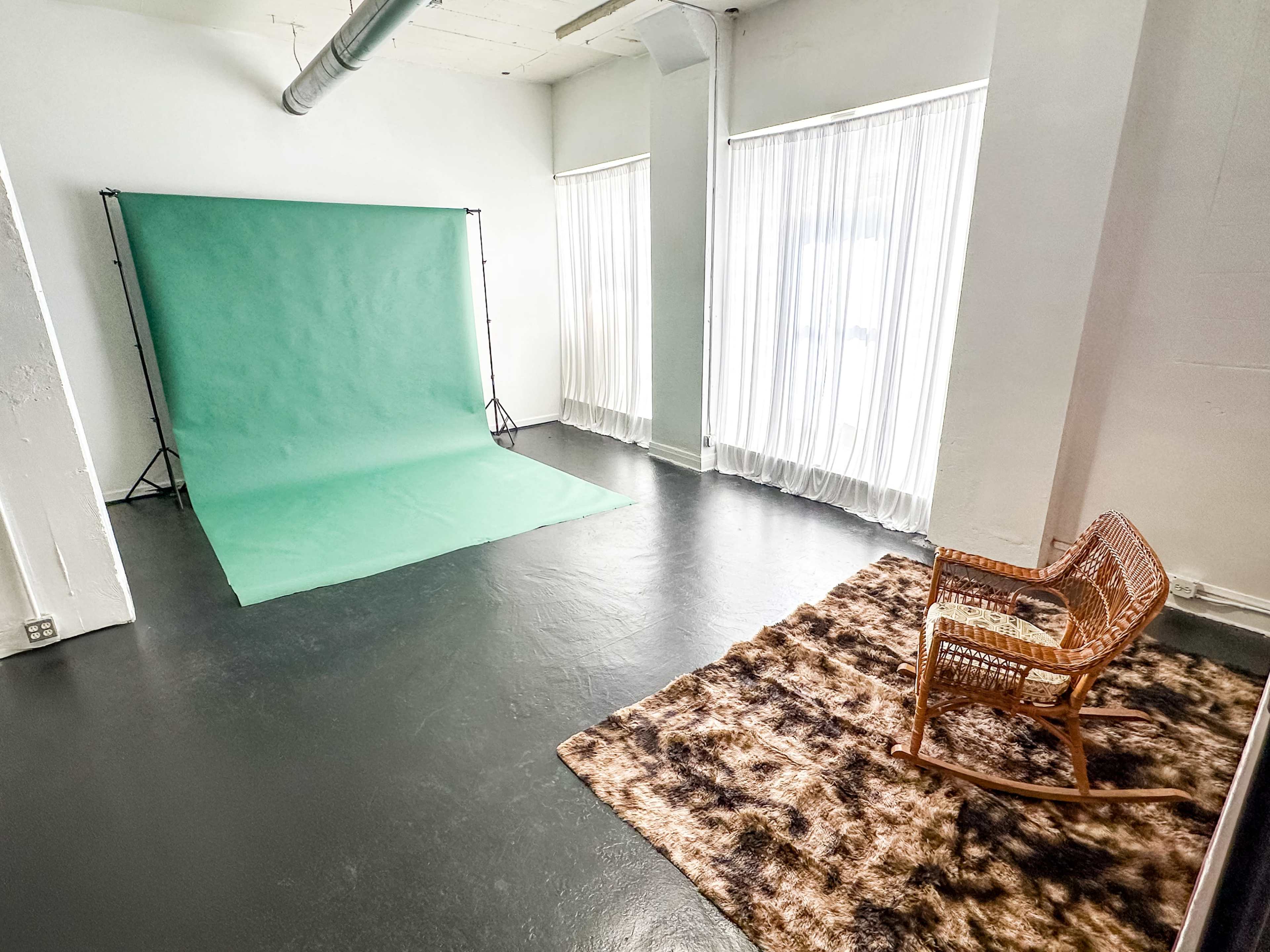 The image shows a bright studio space with a green backdrop, large windows covered with sheer curtains, a textured rug, and a wicker rocking chair.