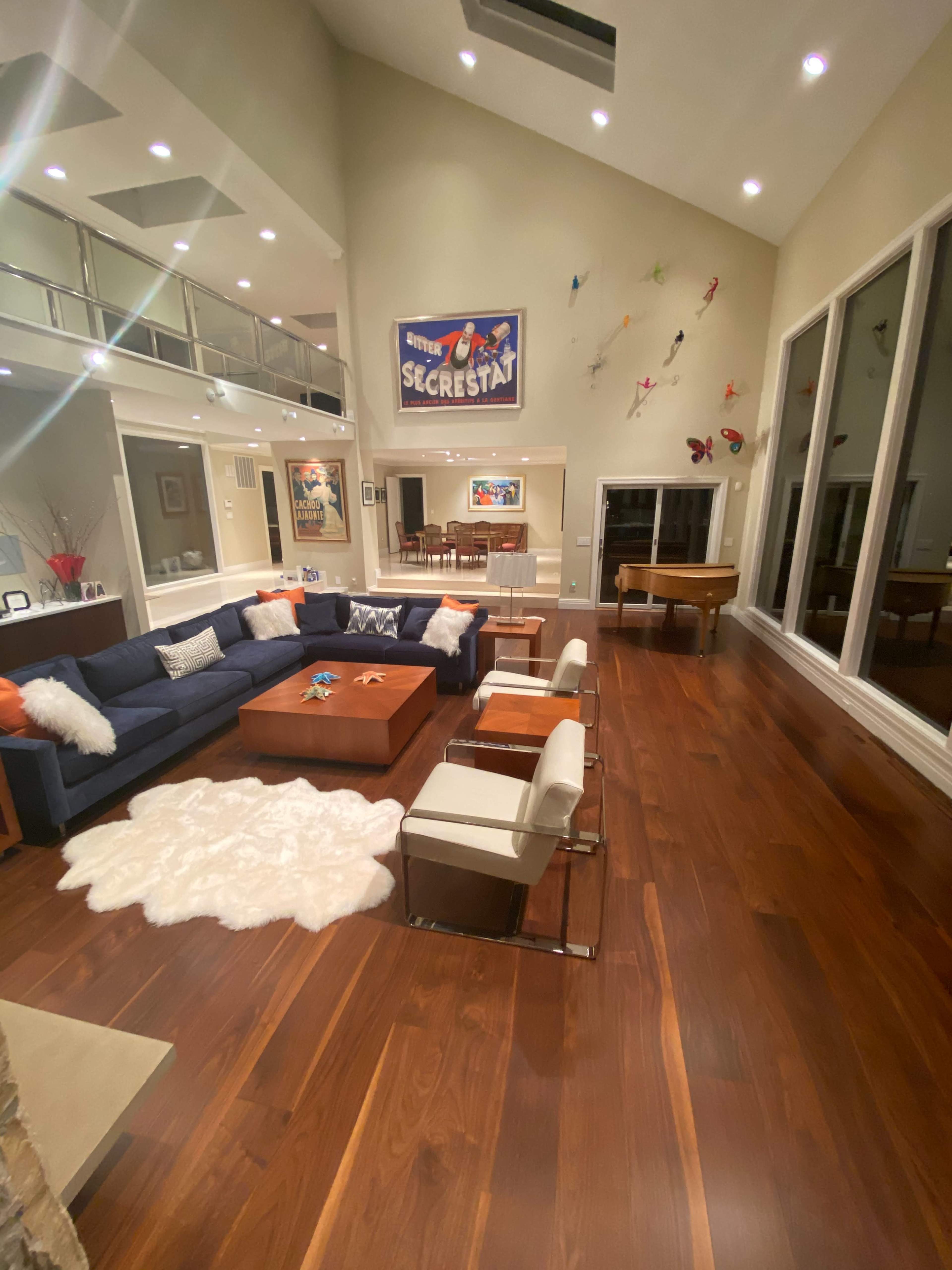 The spacious living room features a blue sectional sofa, a wooden coffee table, and large windows overlooking a dining area and a piano.