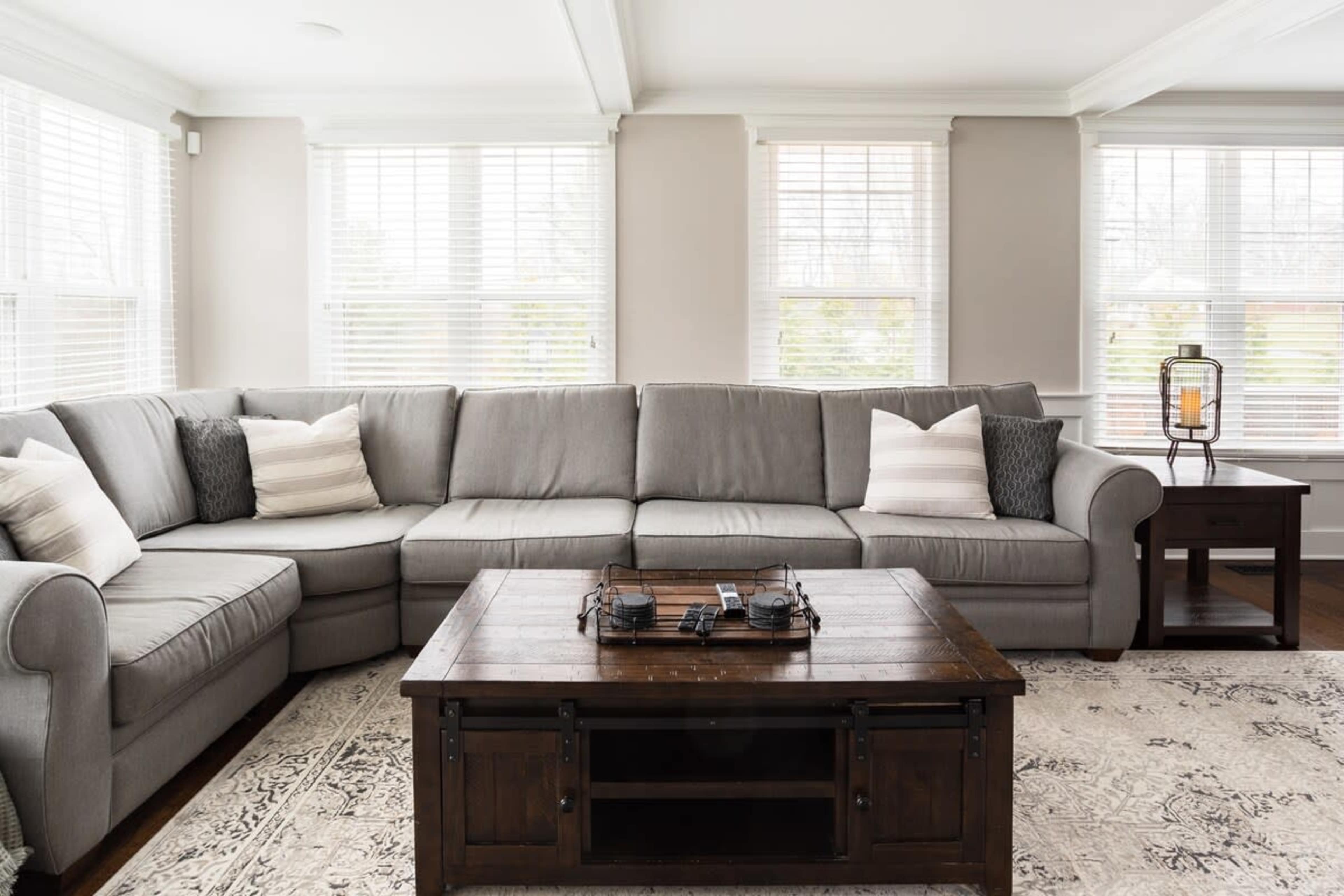 A light gray sectional sofa with decorative pillows, positioned around a dark wooden coffee table on a patterned rug, in a room filled with natural light from multiple windows.
