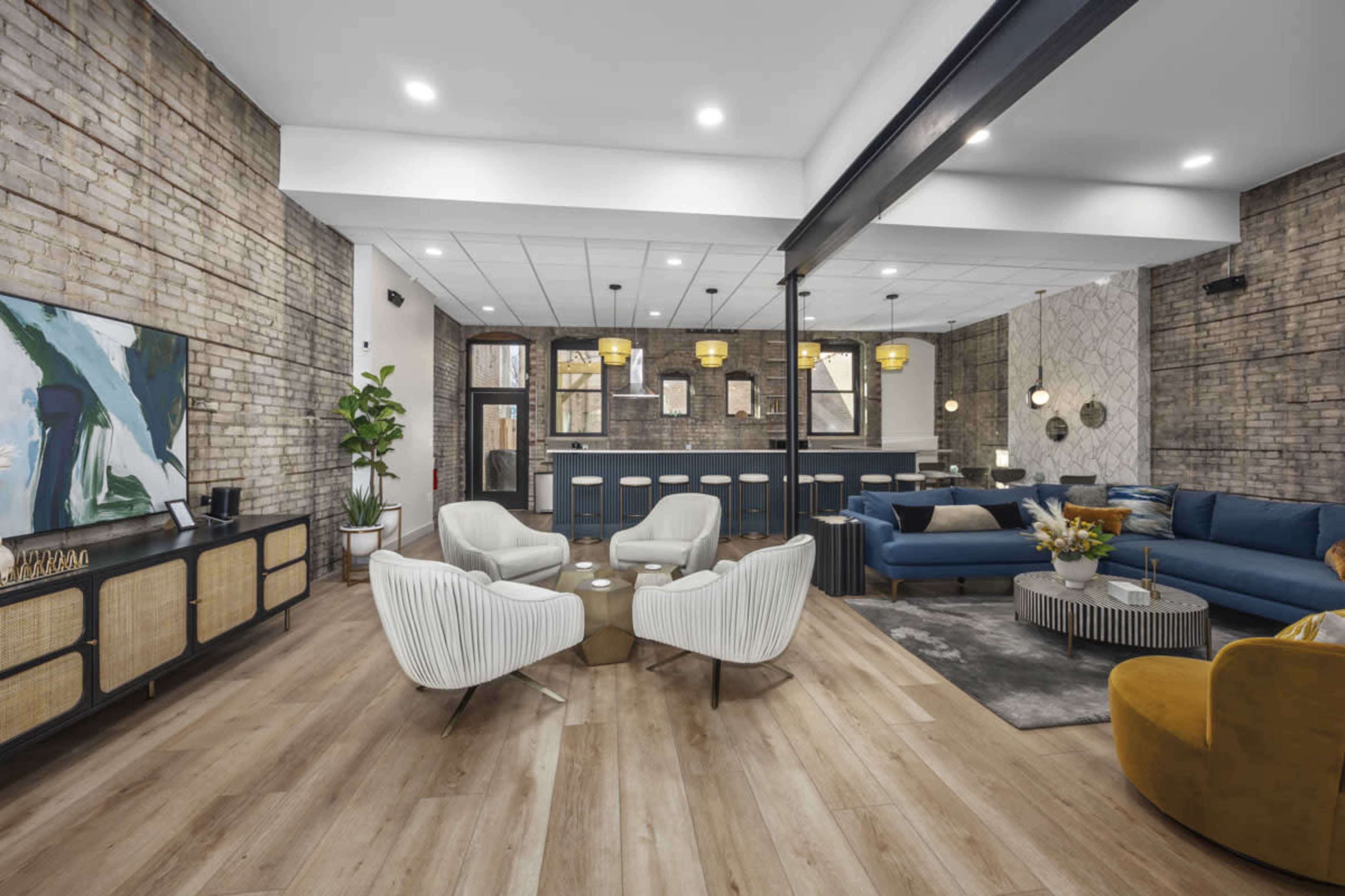 A modern lounge area features a mix of seating options, including white armchairs, a blue sofa, and a yellow accent chair, arranged around a coffee table and adorned with decorative elements against a backdrop of textured brick walls.