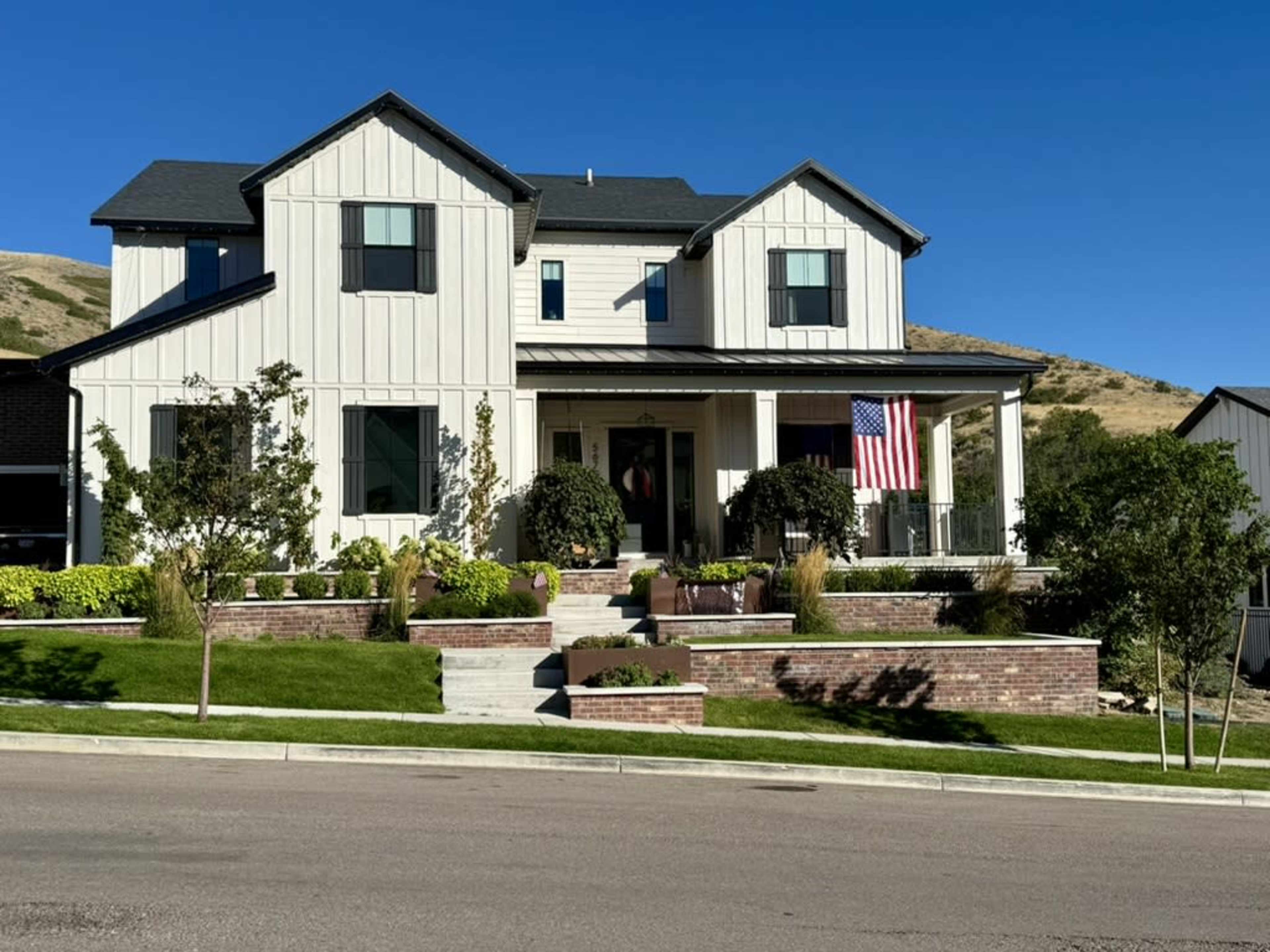 A two-story white house with a black roof features a front porch, landscaped gardens, and an American flag displayed.