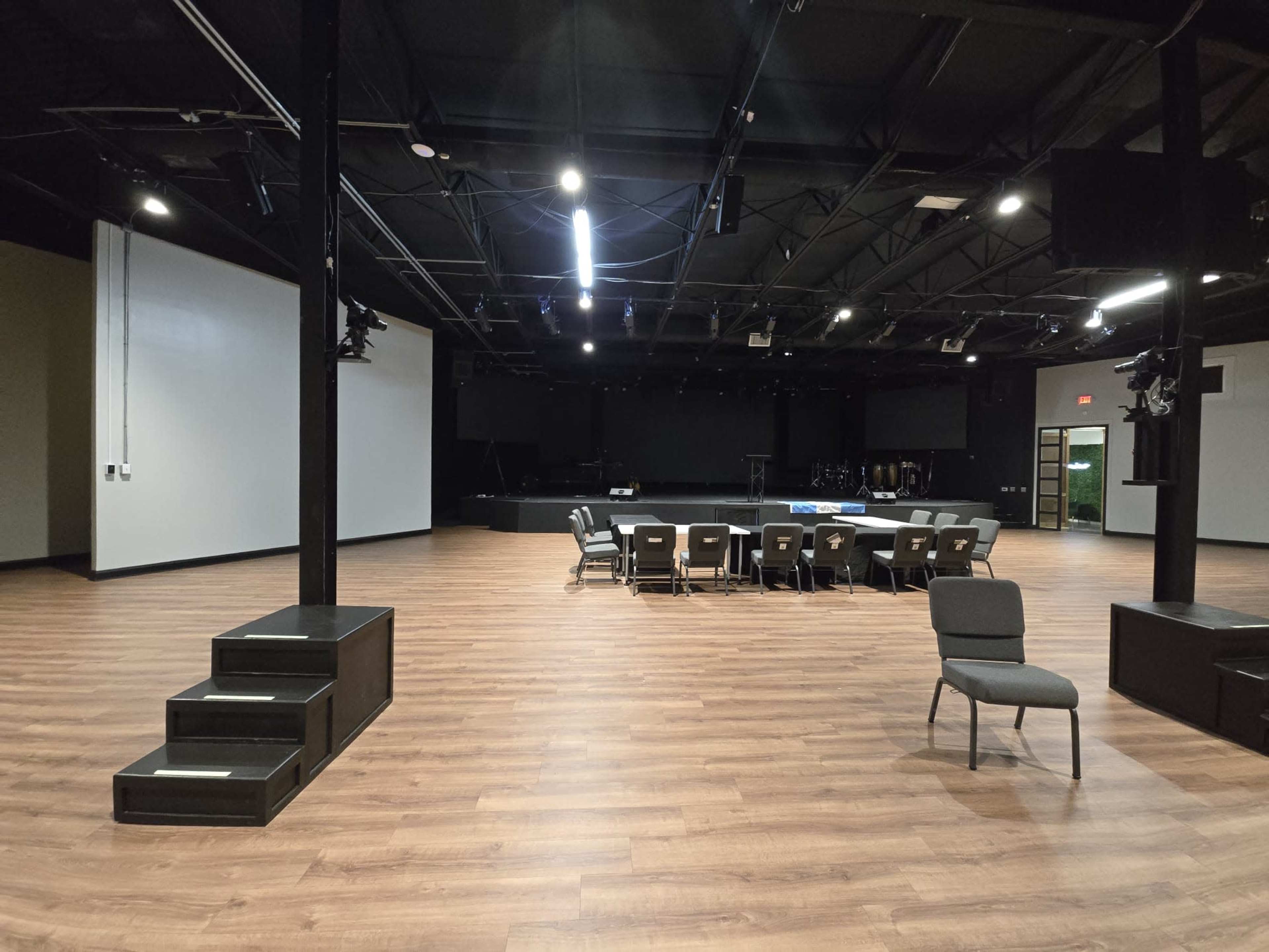 The image shows a spacious indoor venue with wooden flooring, featuring a large open area, a set of steps, a circular arrangement of chairs, and a stage at the back.