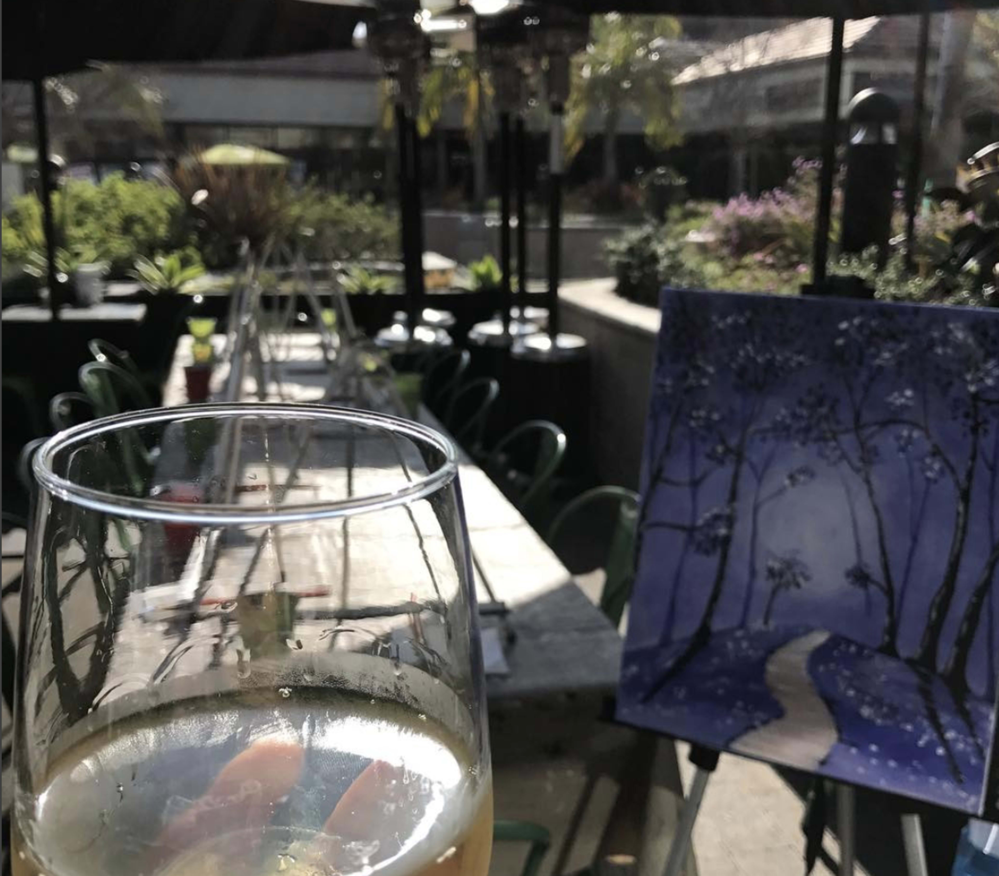 A glass of beverage in the foreground with a painted canvas of a snowy path and trees in the background, set in an outdoor dining area.