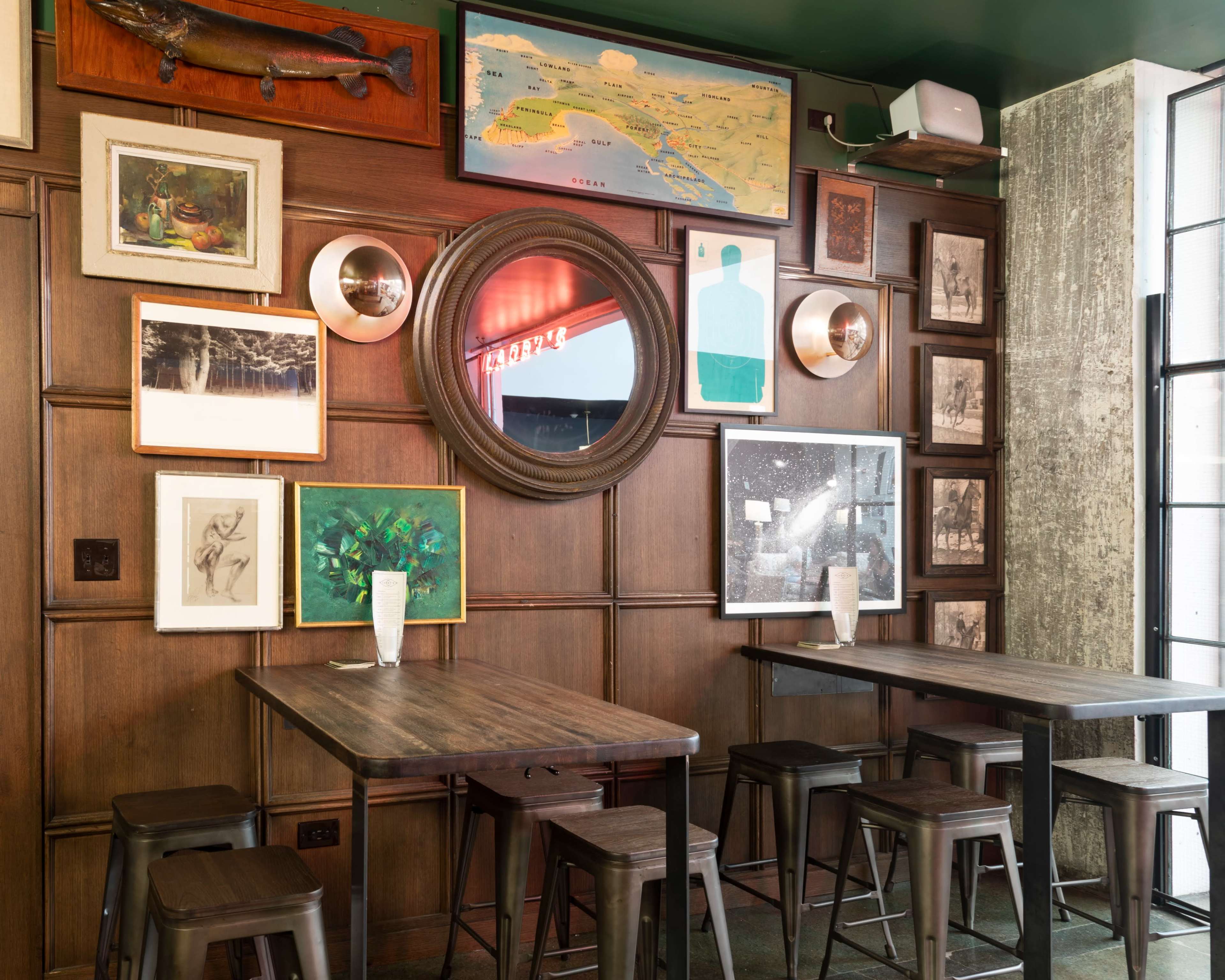 The image showcases a wooden-paneled wall adorned with various framed artworks and mirrors, accompanied by two minimalist metal tables and stools in a stylish interior setting.