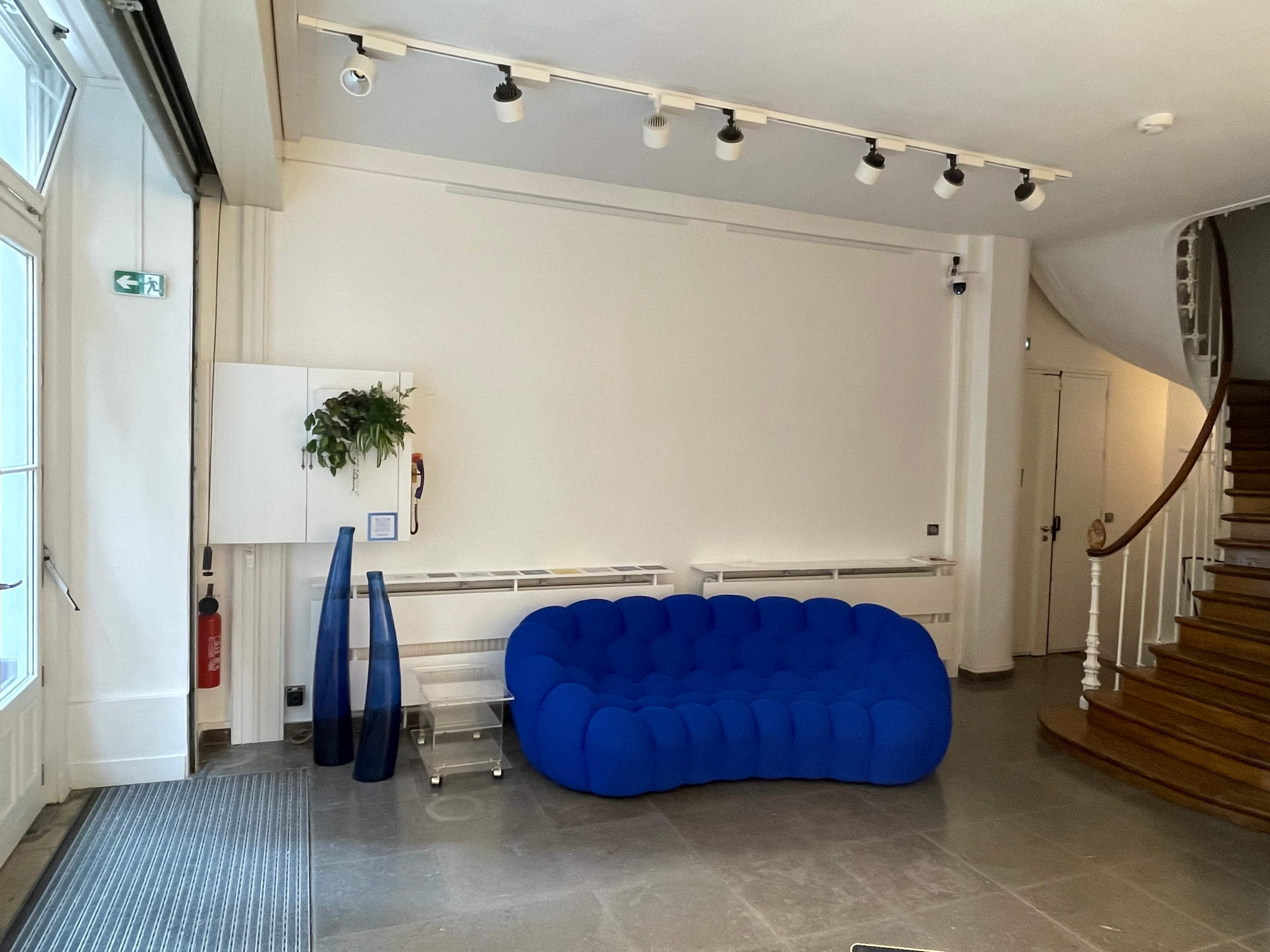 A modern interior space featuring a blue, sculptural couch, a small potted plant on the wall, and a spiral staircase in the background.